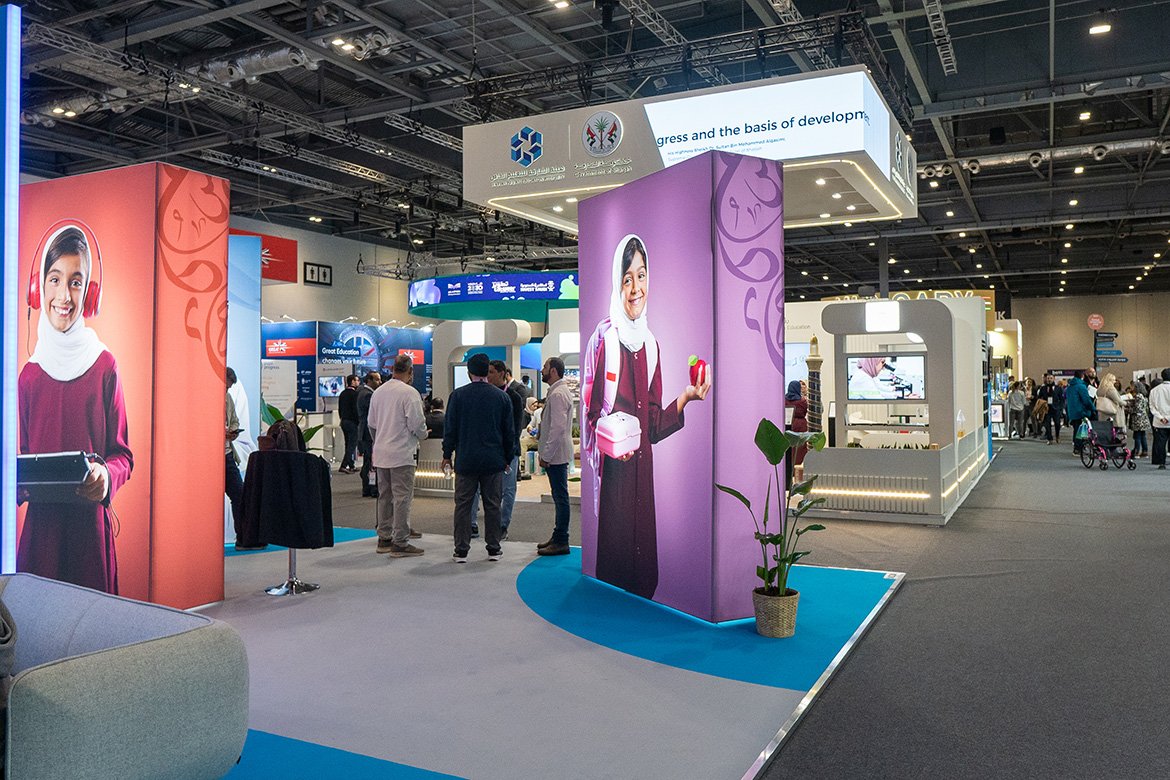 Trade show booth with large promotional images of women, some in hijabs, inside an exhibition hall with people walking around and engaging with displays.