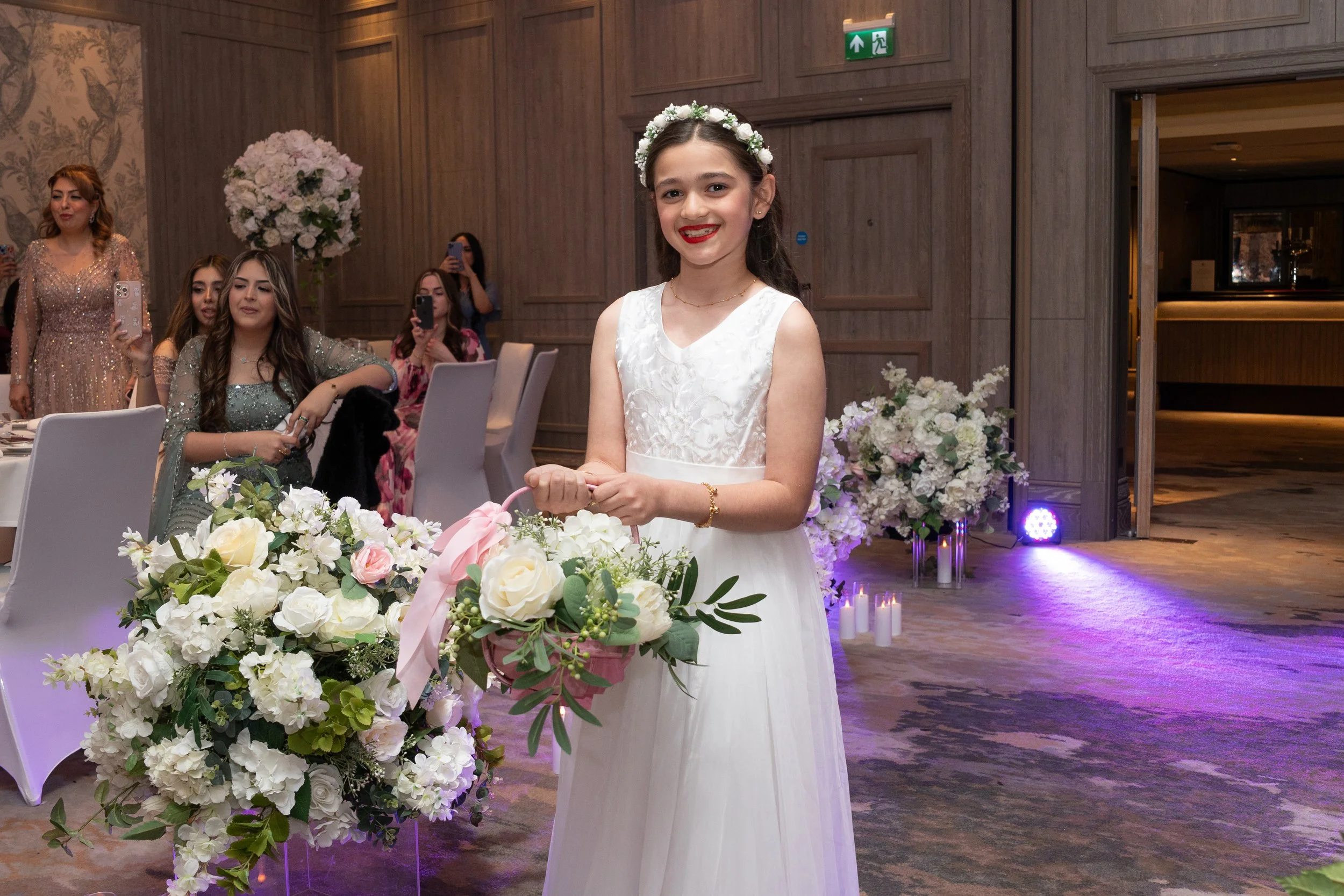 Young girl in a white dress with a floral headband, holding a bouquet of flowers, at a celebration or wedding event.