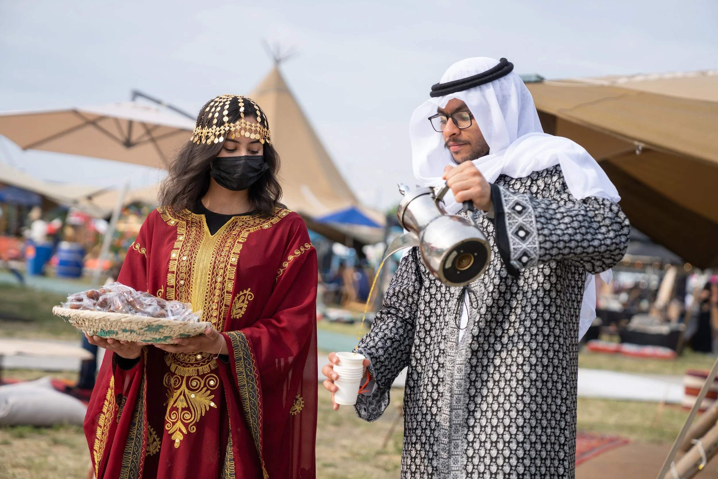 A man and woman dressed in traditional Middle Eastern attire at an outdoor festival. The woman wears a red and gold embroidered dress with a decorative headpiece and black face mask, holding a basket of baked goods. The man wears a patterned robe wit