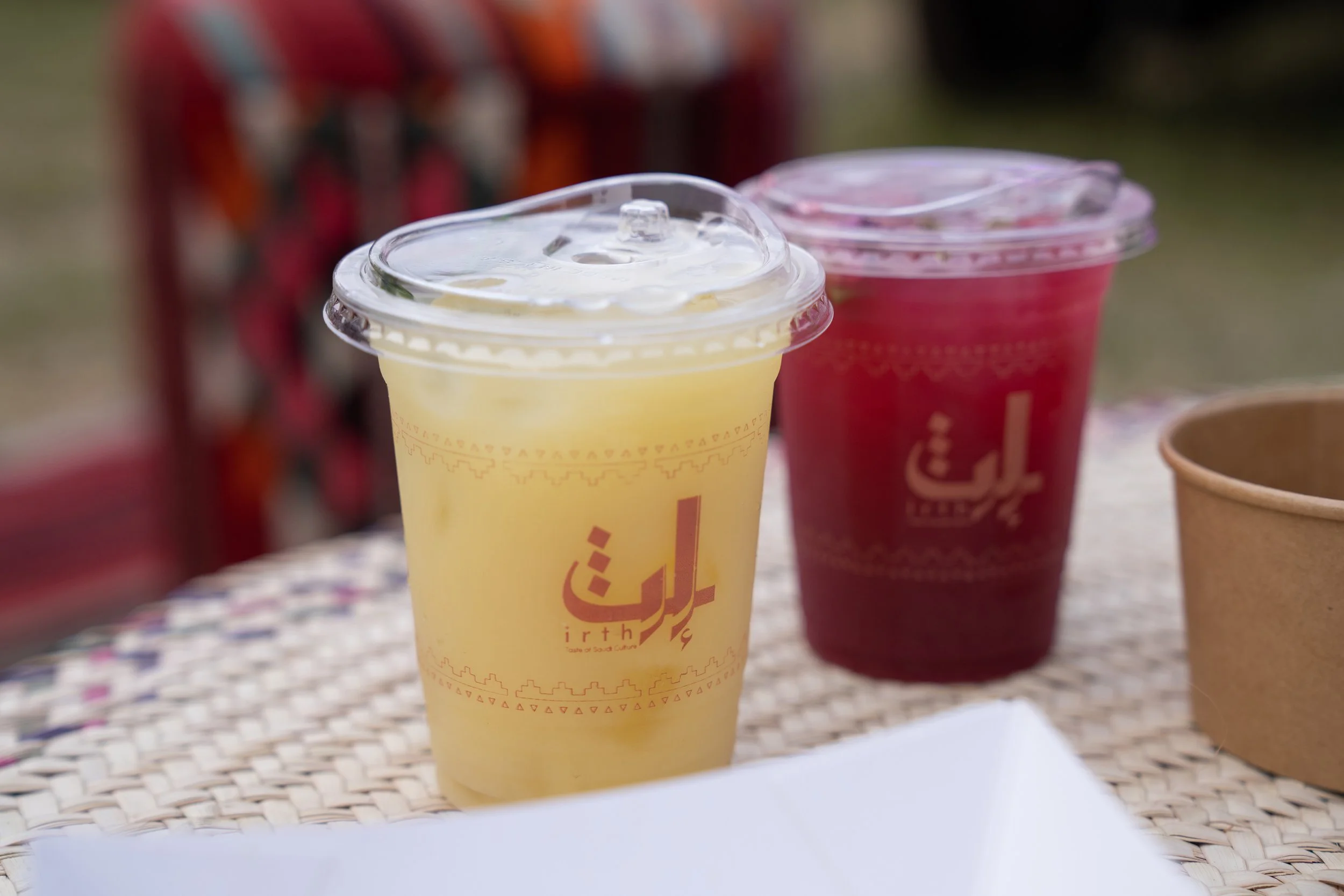 Two plastic cups with lids and straws, one yellow-colored and one red-colored, placed on a woven table surface outdoors. The cups have Arabic writing and the word "irth" in english, likely indicating beverages from a Middle Eastern or Arabic café.