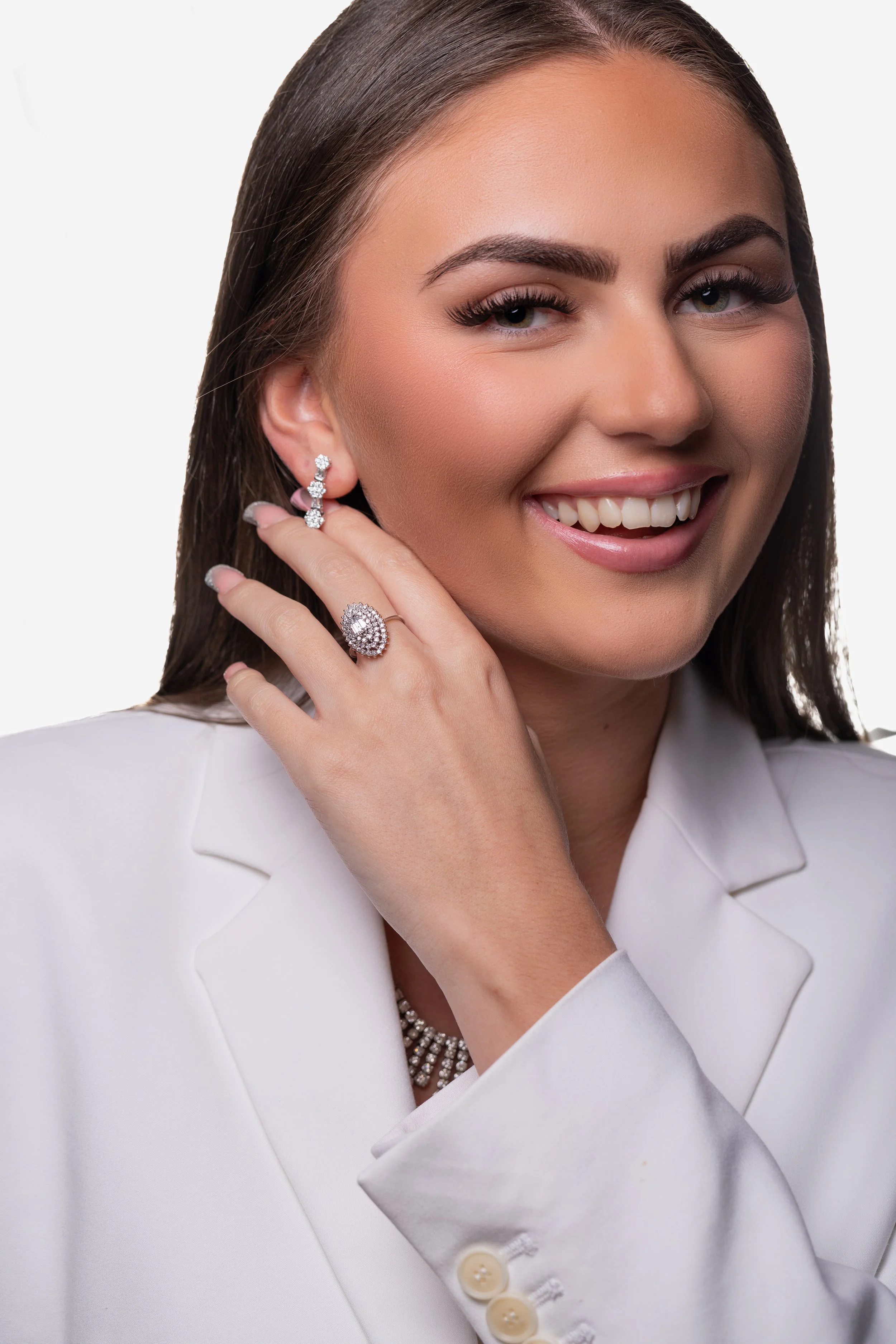 A woman smiling while touching her face, wearing a white blazer, diamond earrings, a ring, and a necklace.