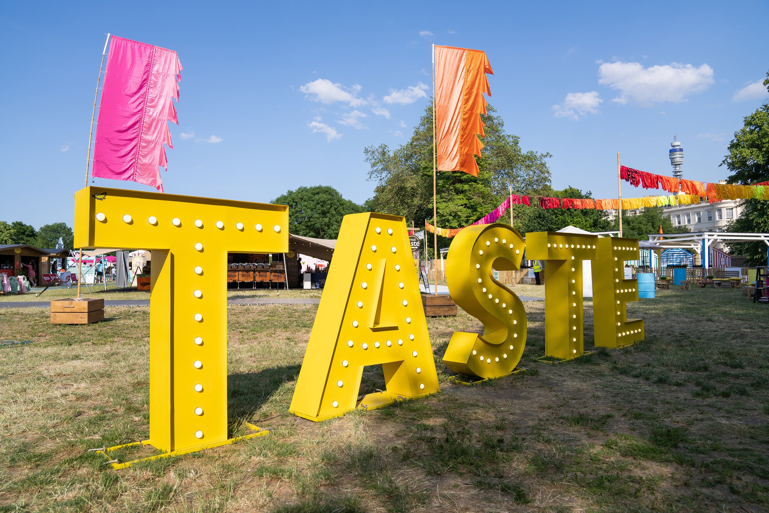 Colorful outdoor festival scene with large yellow marquee letters spelling 'TASTE'. Flags and banners are hanging in the background, and tents and booths are set up on the grassy area under a blue sky.