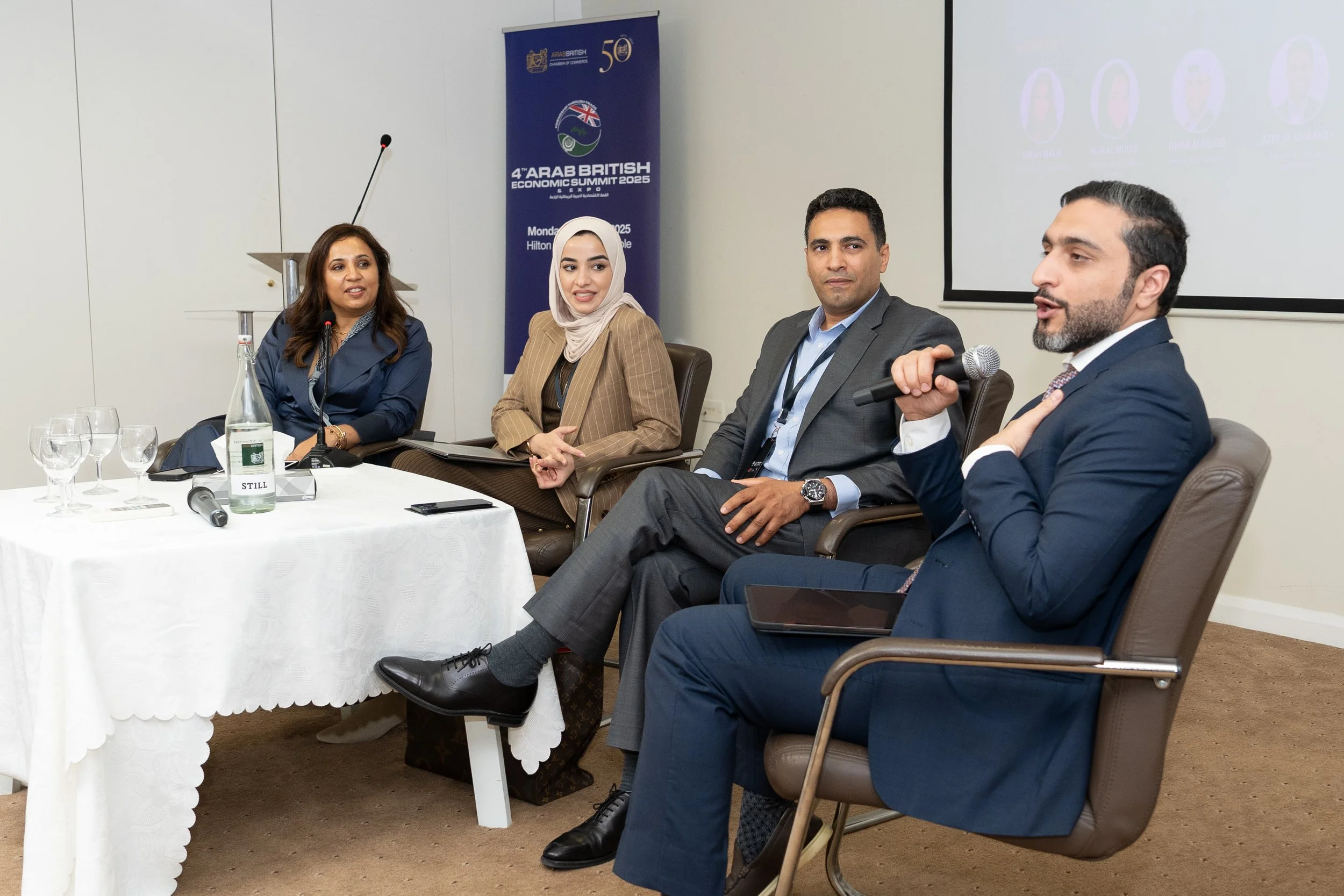 Four professionals sitting on a panel at a conference, with a blue banner and projection screen behind them. One man appears to be speaking into a microphone.