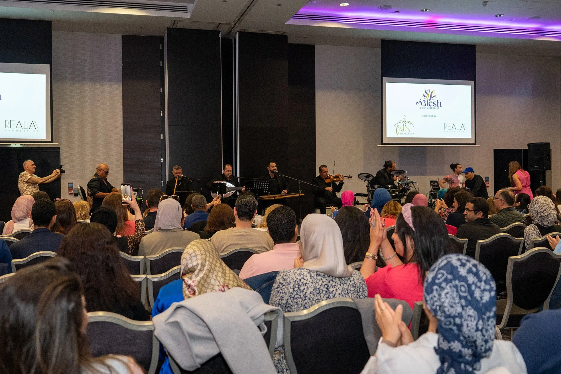 Audience attending a live musical performance in a conference room with musicians playing instruments on stage and two screens displaying event logos.