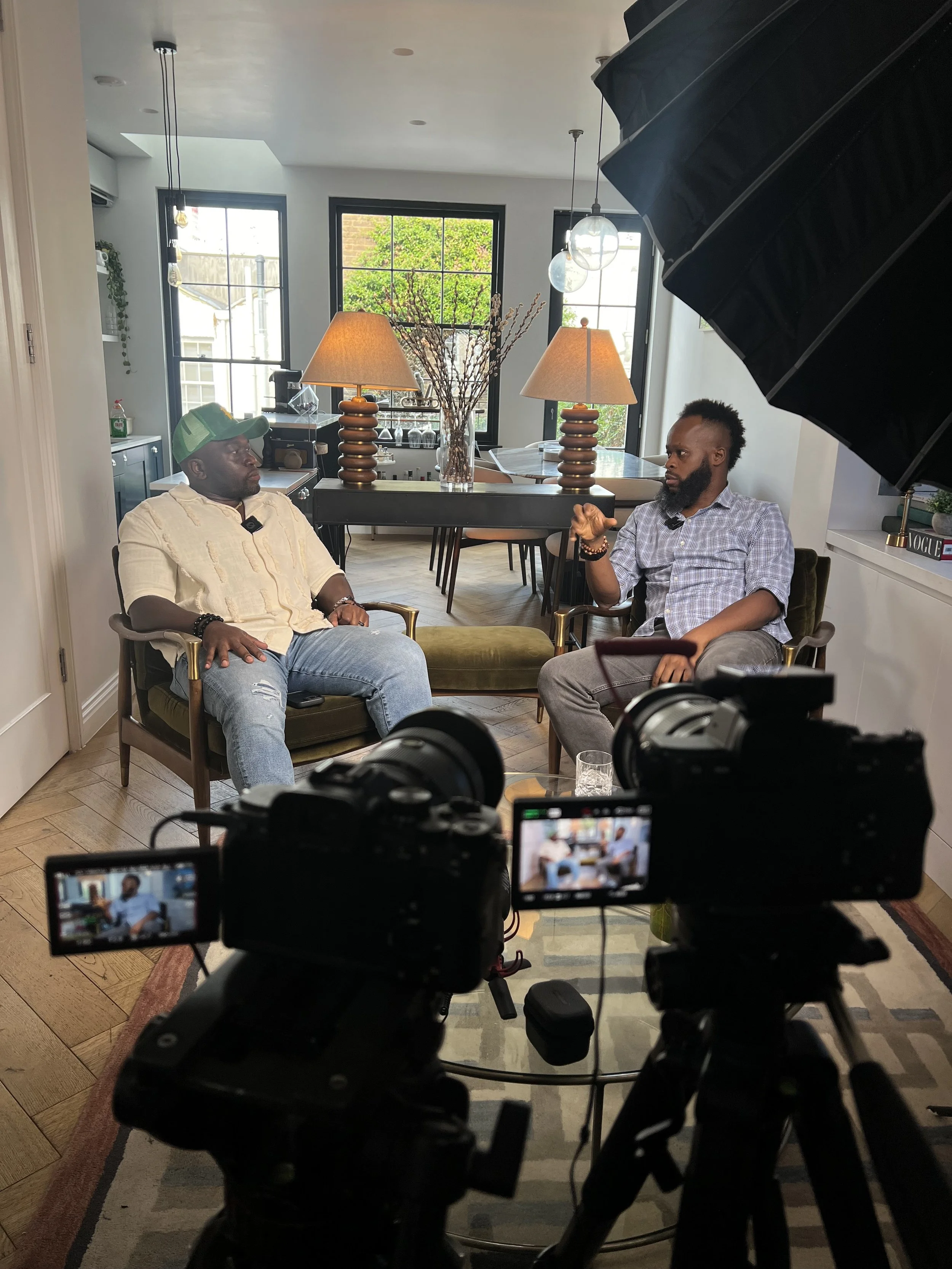 Two men are sitting and talking in a home studio or interview setting, with a camera recording them. The room is well-lit with natural light from large windows, and has a modern decor with lamps and a dining table in the background.