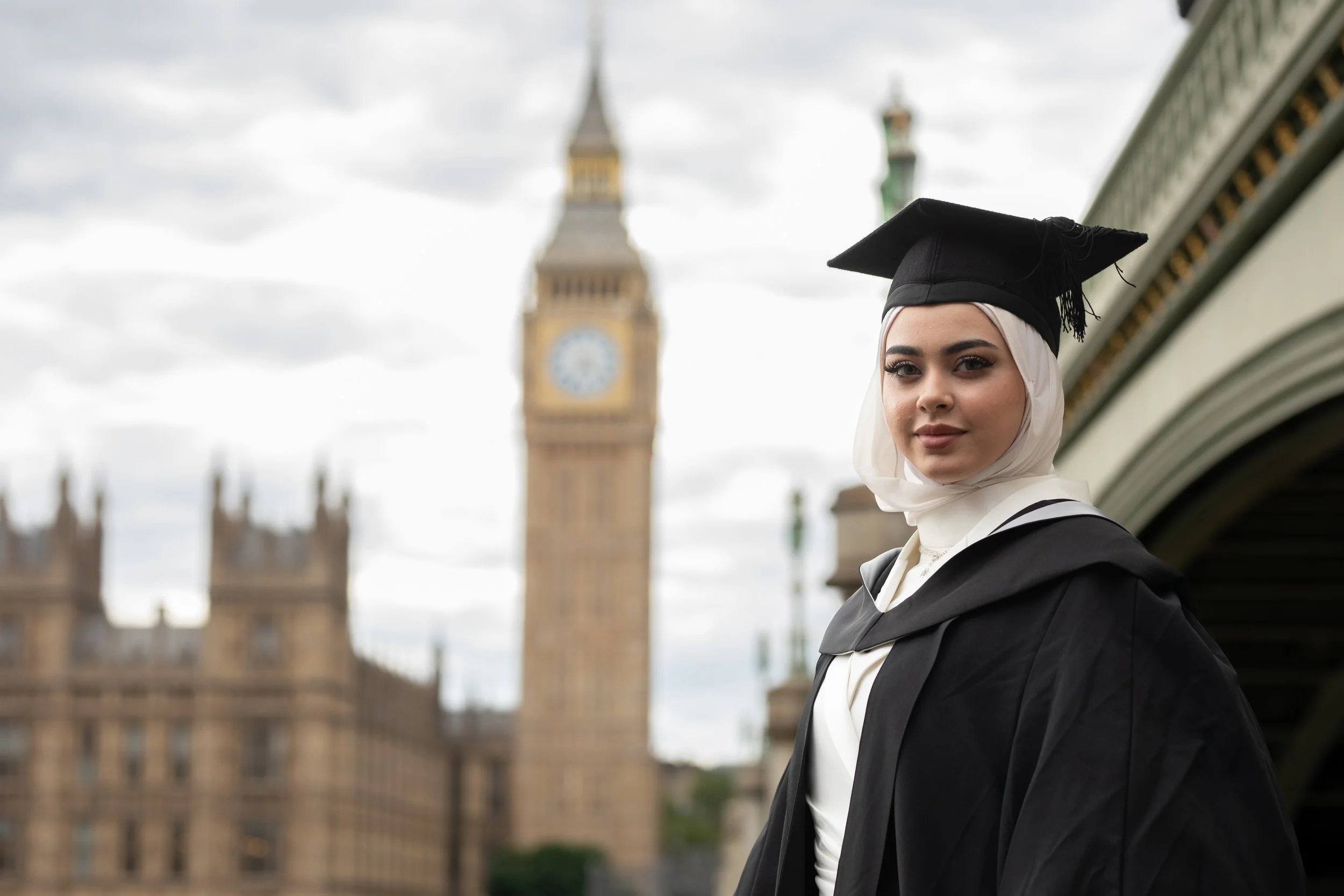 A young woman in graduation attire, wearing a black cap and gown, standing outdoors with Big Ben and the Houses of Parliament in London in the background.
