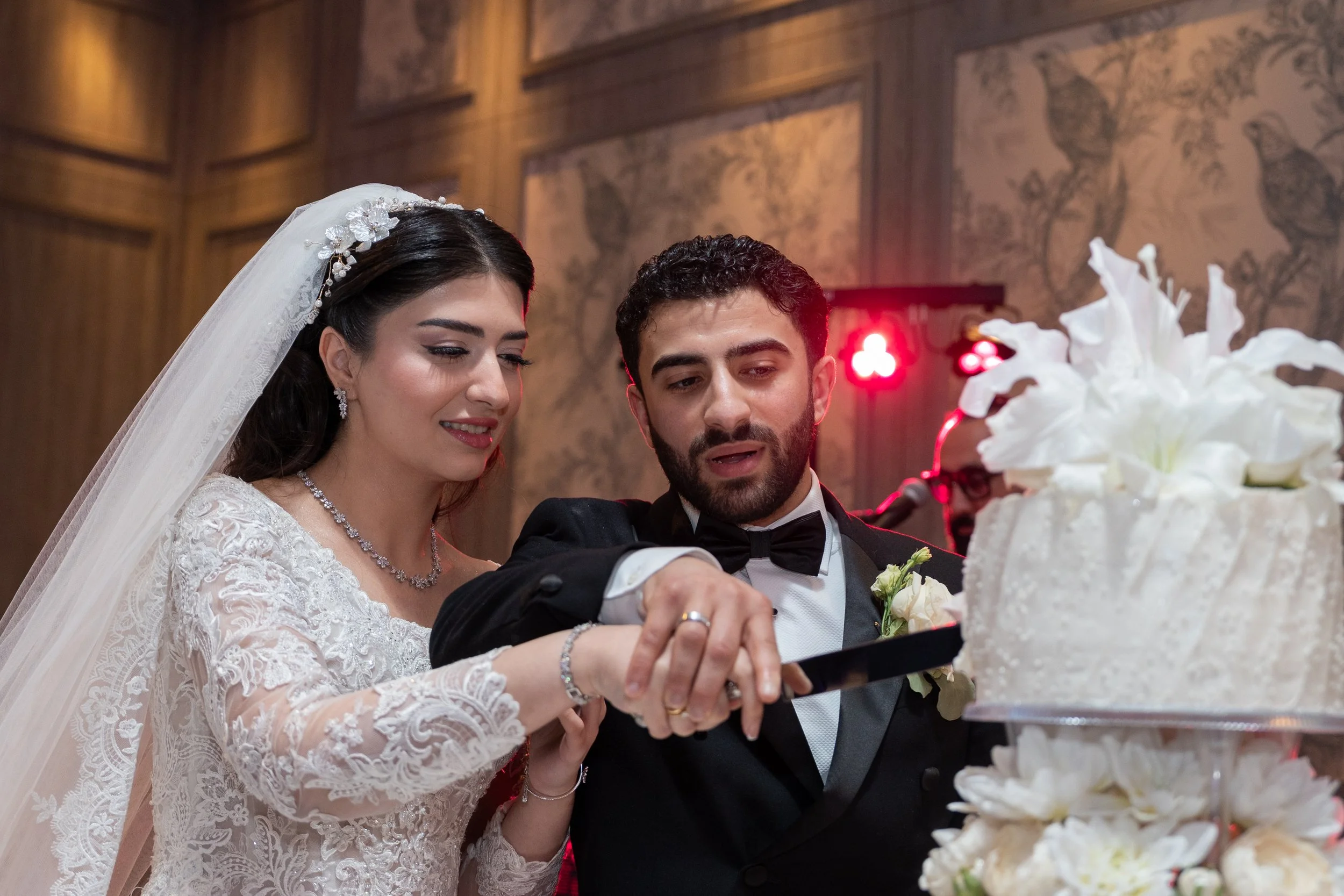 Bride and groom cutting wedding cake in a decorated venue with wood-paneled walls and floral artwork.