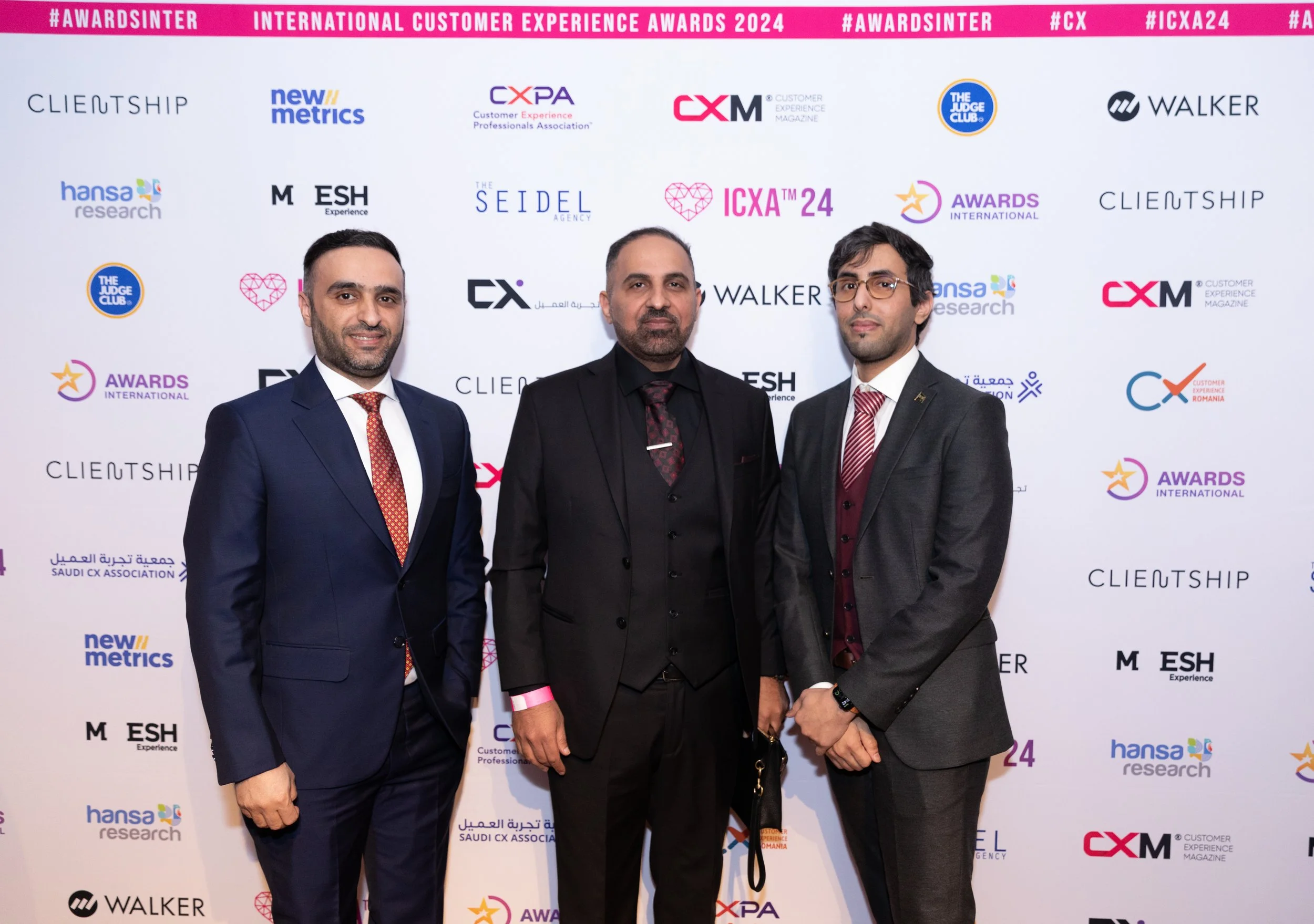 Three men in formal suits standing in front of a backdrop with event and sponsor logos at the International Customer Experience Awards 2024, smiling and posing for the photo.