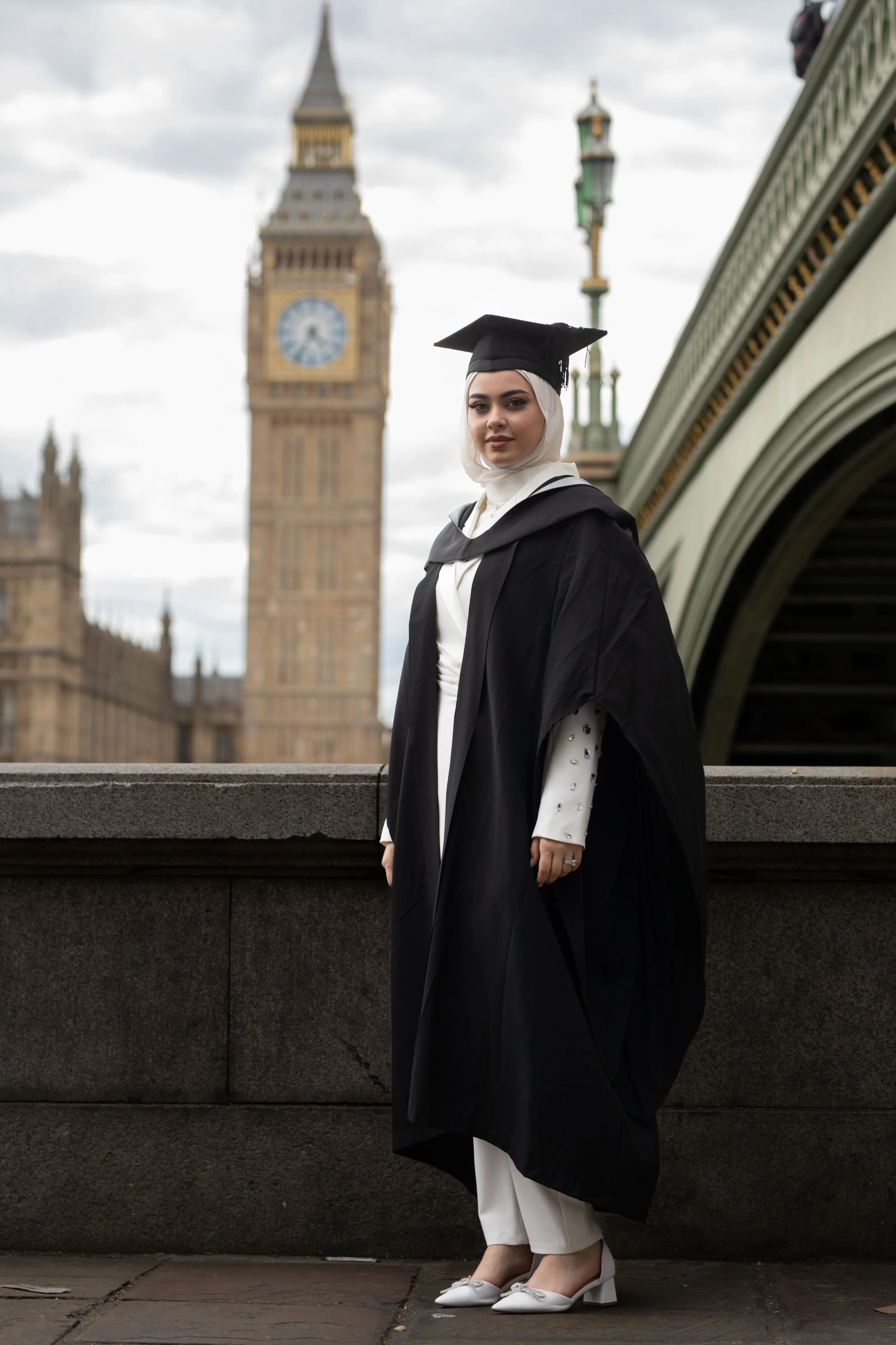 A woman in a graduation cap and gown standing outdoors near Big Ben in London.