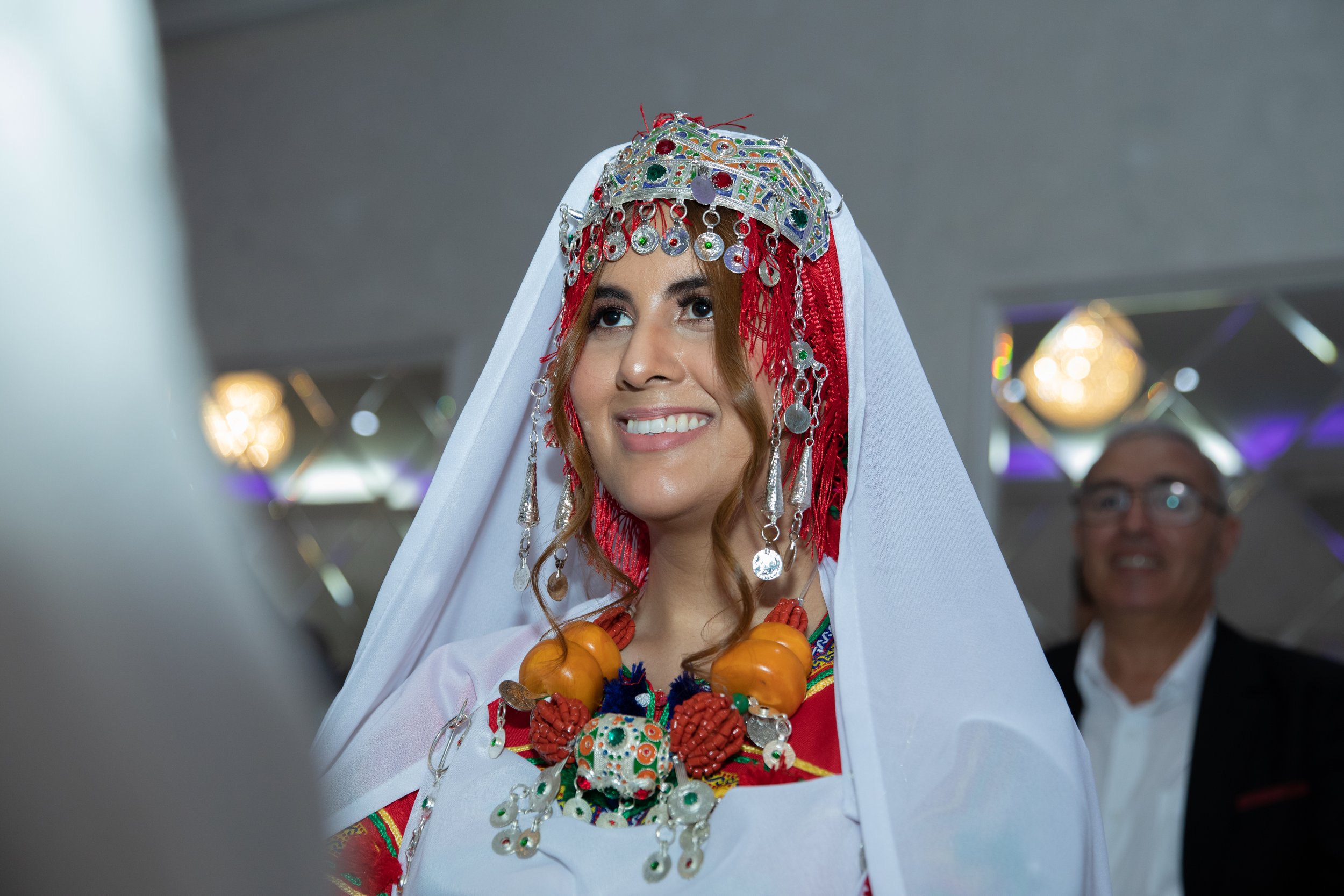 Woman dressed in traditional clothing with jewelry, a white veil, and a necklace made of fruits, smiling in an indoor setting.