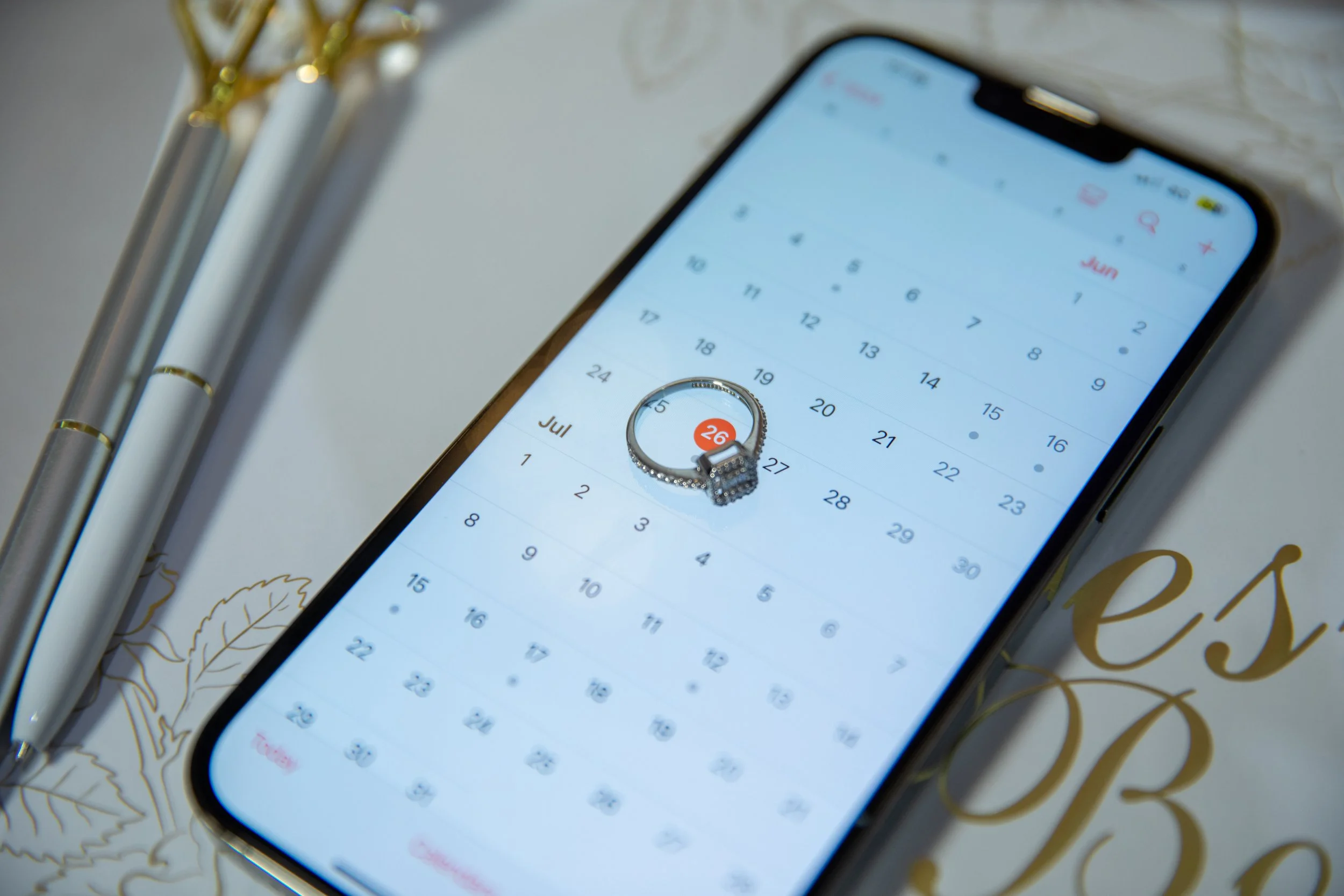 Smartphone displaying a calendar for June and July with a marked date of June 26, placed on a decorative surface with a ring on top of the screen and adjacent pens.