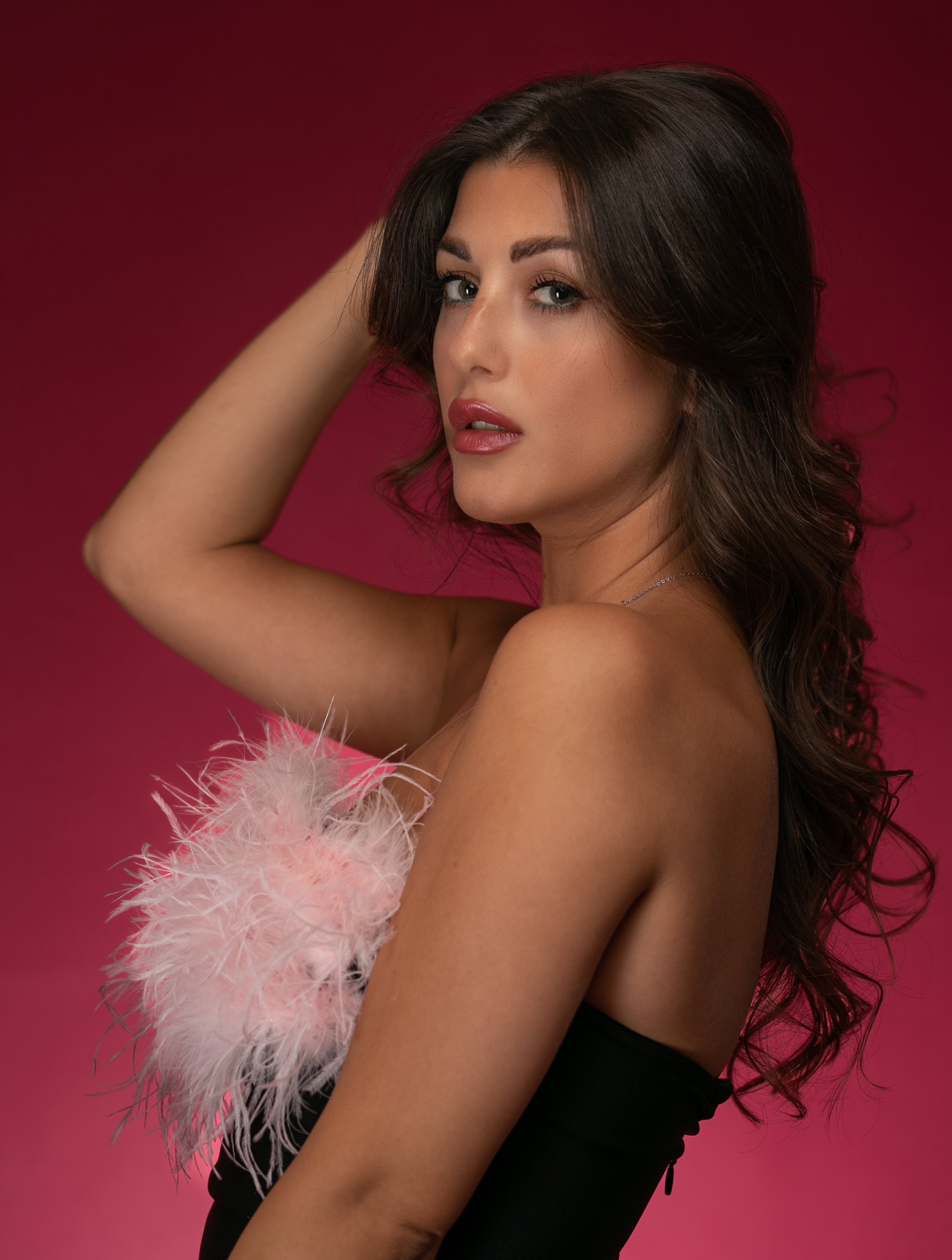 A young woman with dark brown styled hair, wearing a black strapless outfit with pink feather accents, posing with her arm raised and looking at the camera against a pink background.