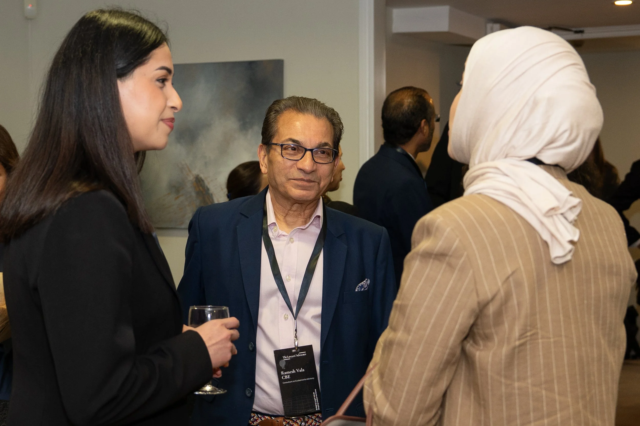 Three people engaged in conversation at a professional networking event, with others in the background.