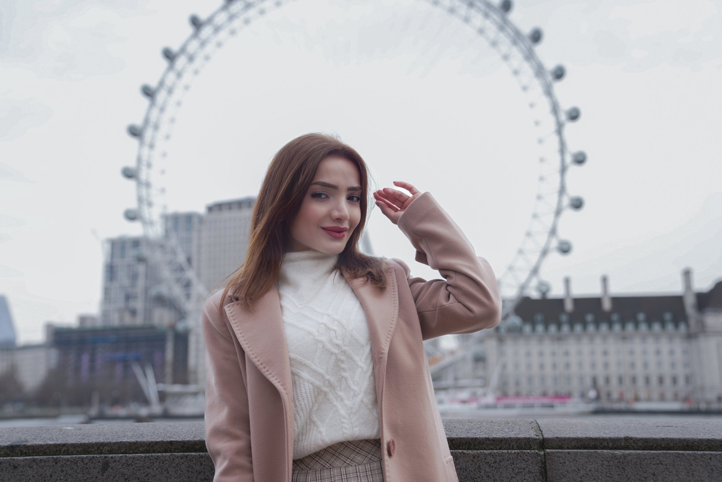 A young woman with brown hair, wearing a beige coat and white sweater, poses near the London Eye Ferris wheel with buildings in the background on a cloudy day.
