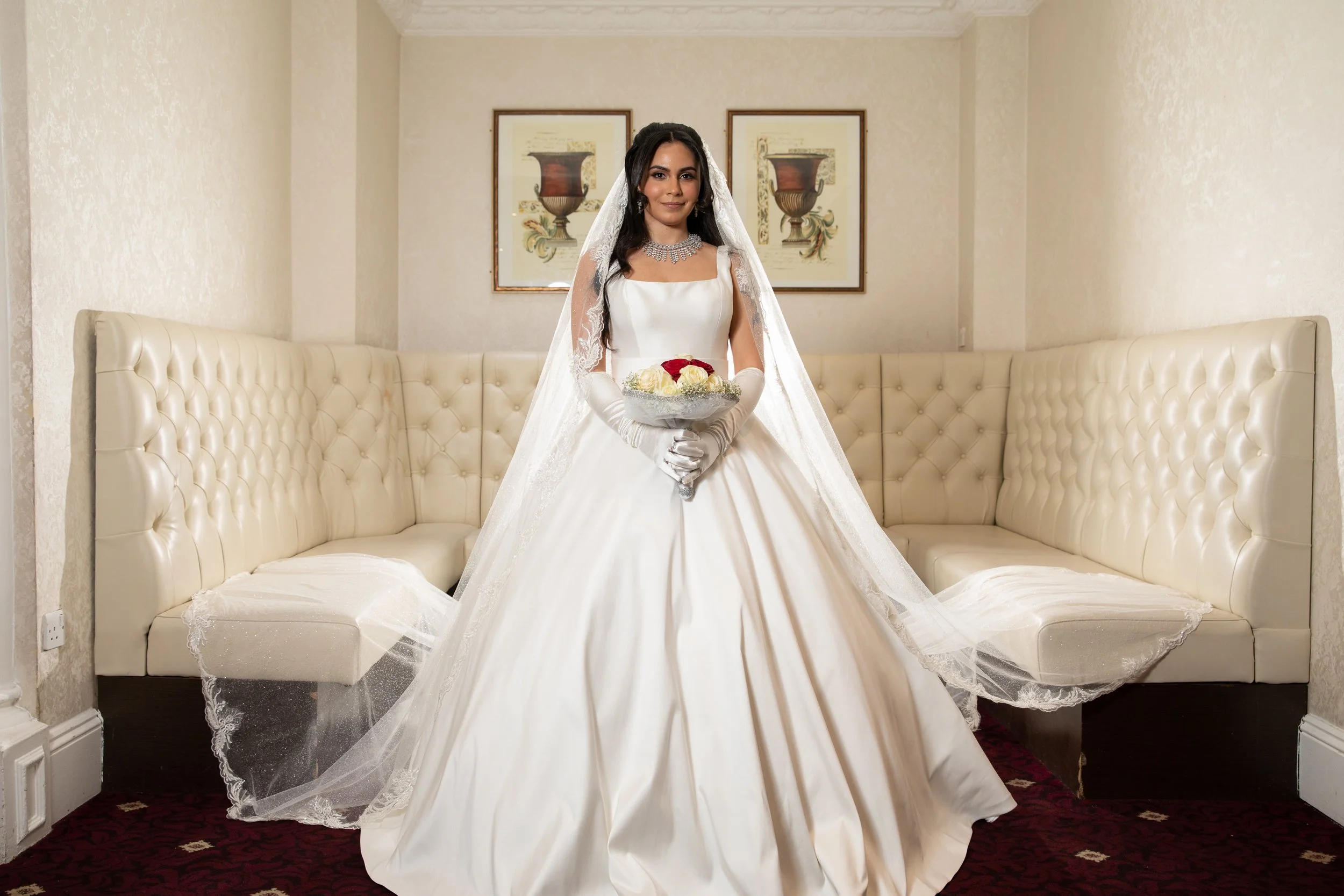 A bride in a white wedding gown, holding a bouquet of flowers, standing in a room with cream-colored sofa and framed artwork on the wall.