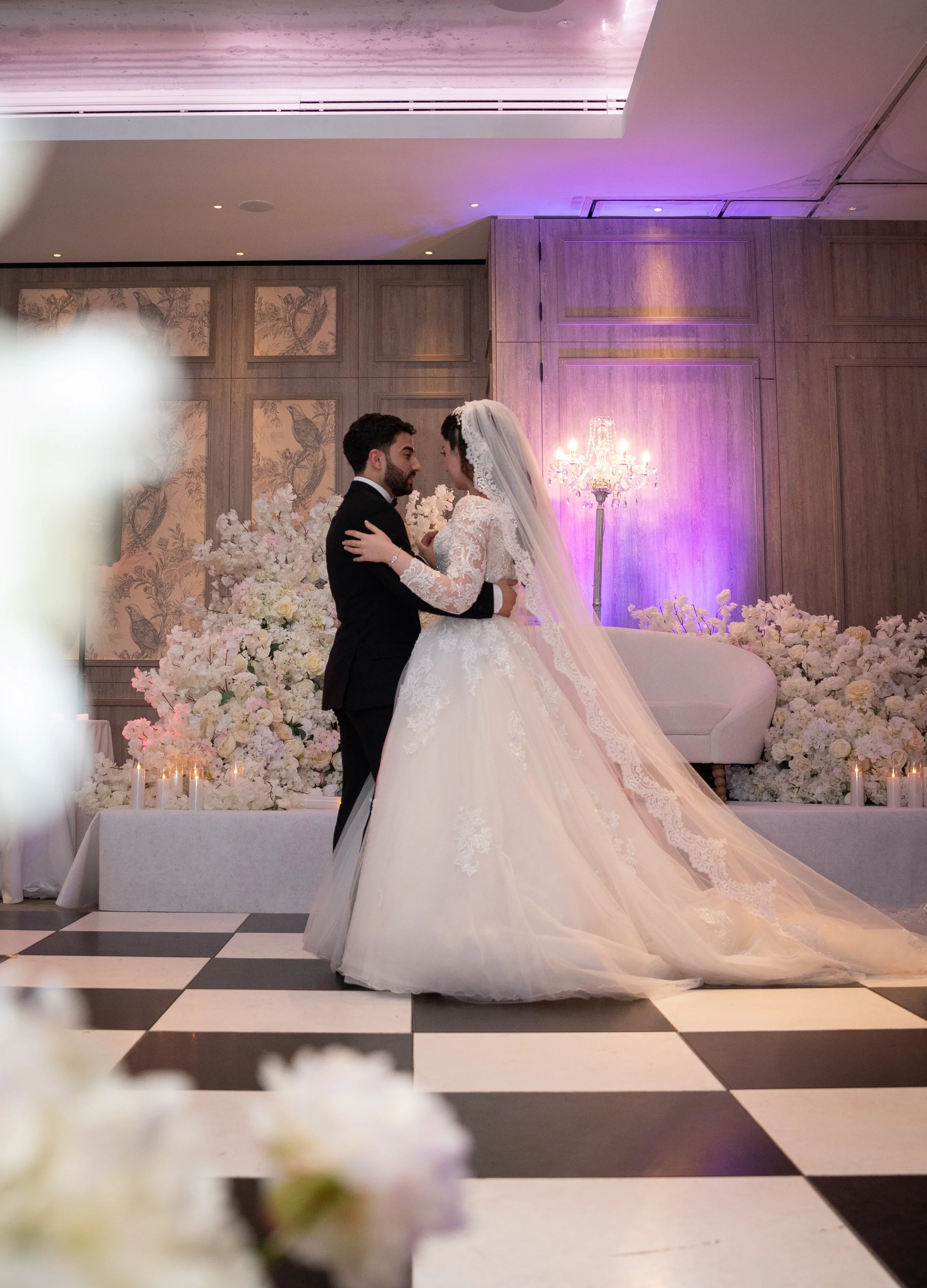A bride and groom dancing at their wedding reception in a decorated ballroom with flowers and soft lighting.