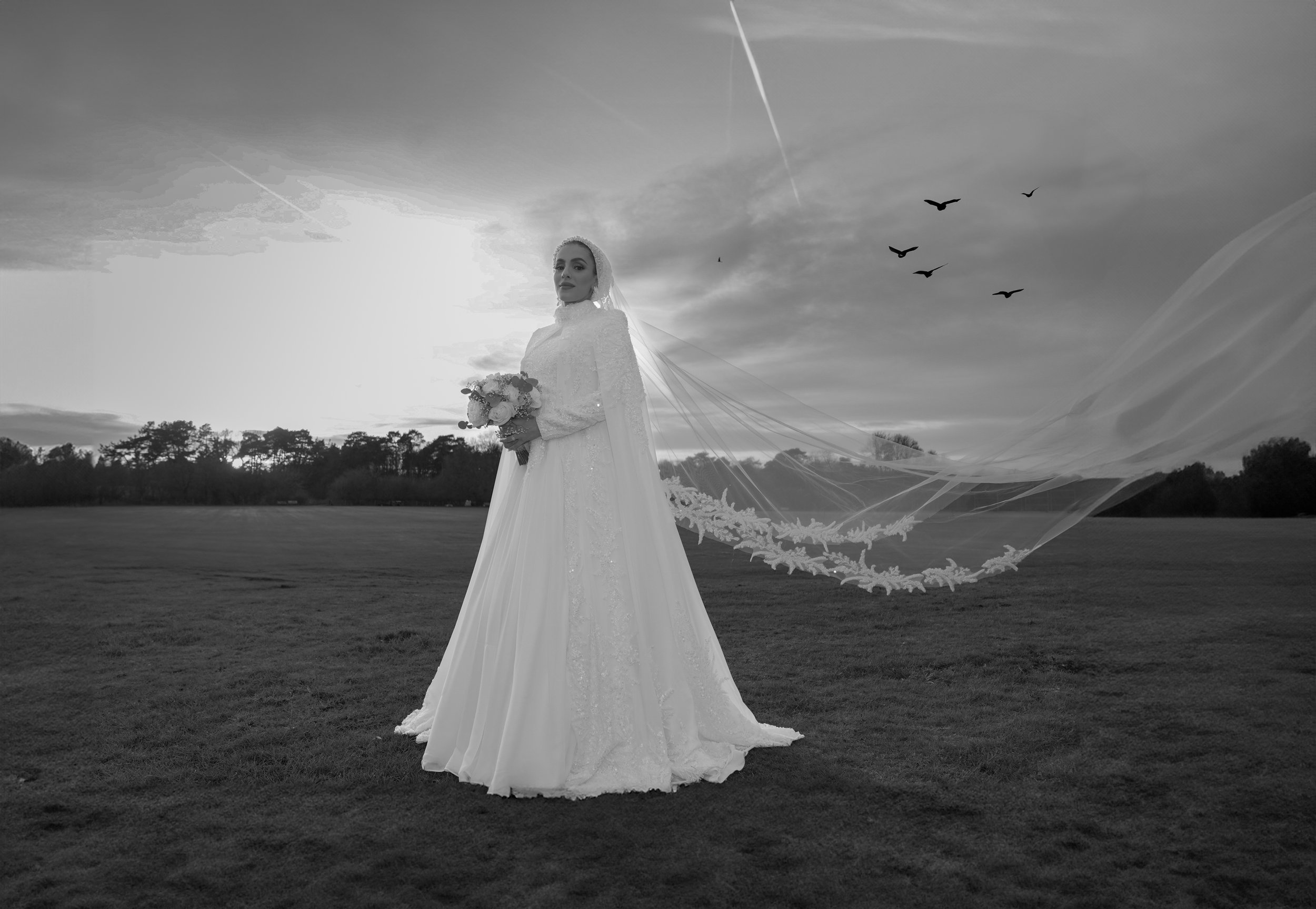 A bride in a long wedding dress holding a bouquet, standing in an open field at sunset with birds flying in the sky.