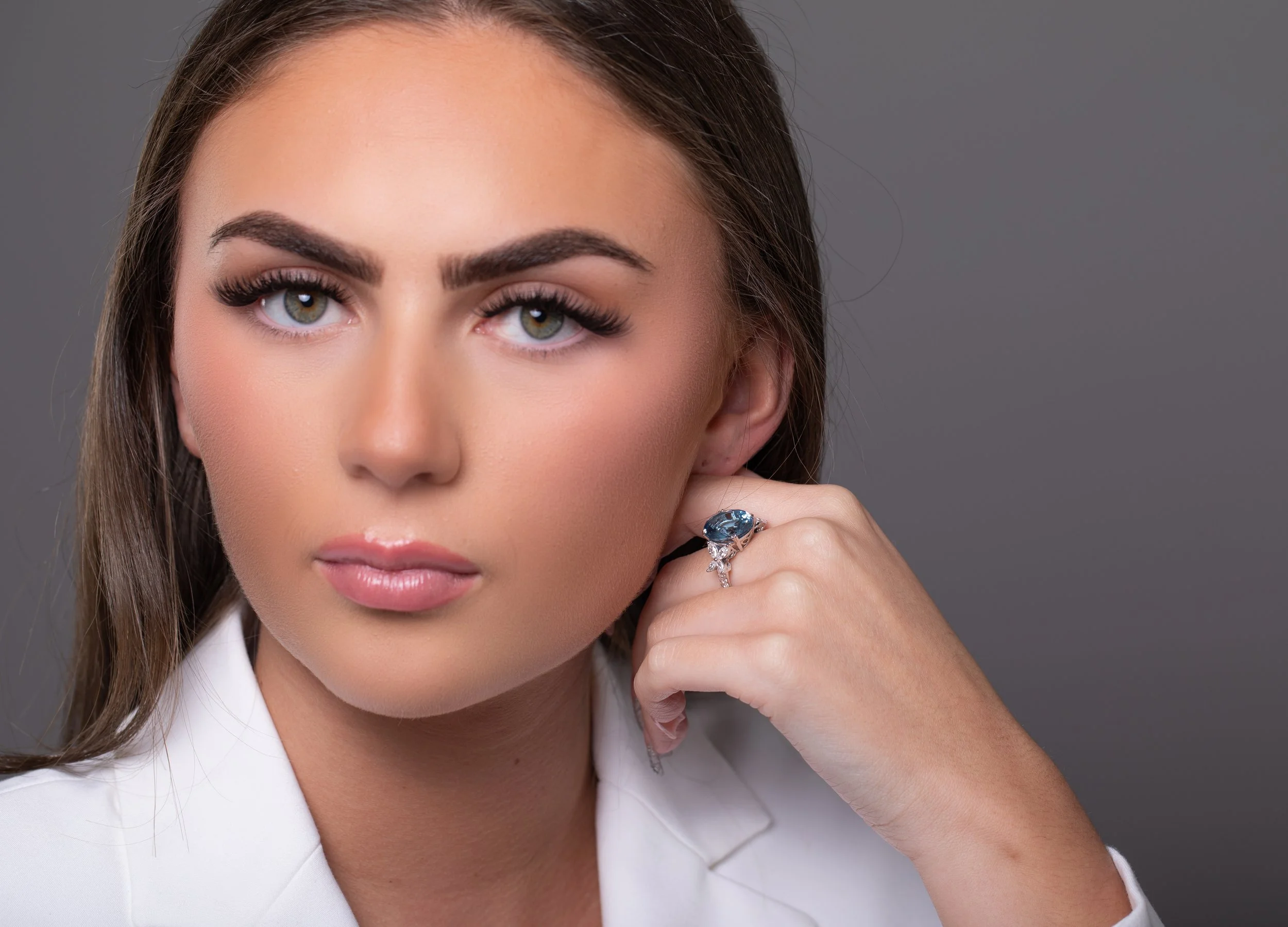 A young woman with long brown hair, light makeup, and a white blazer posing with her hand resting near her face, wearing a ring with a large blue gemstone and small diamonds, against a gray background.