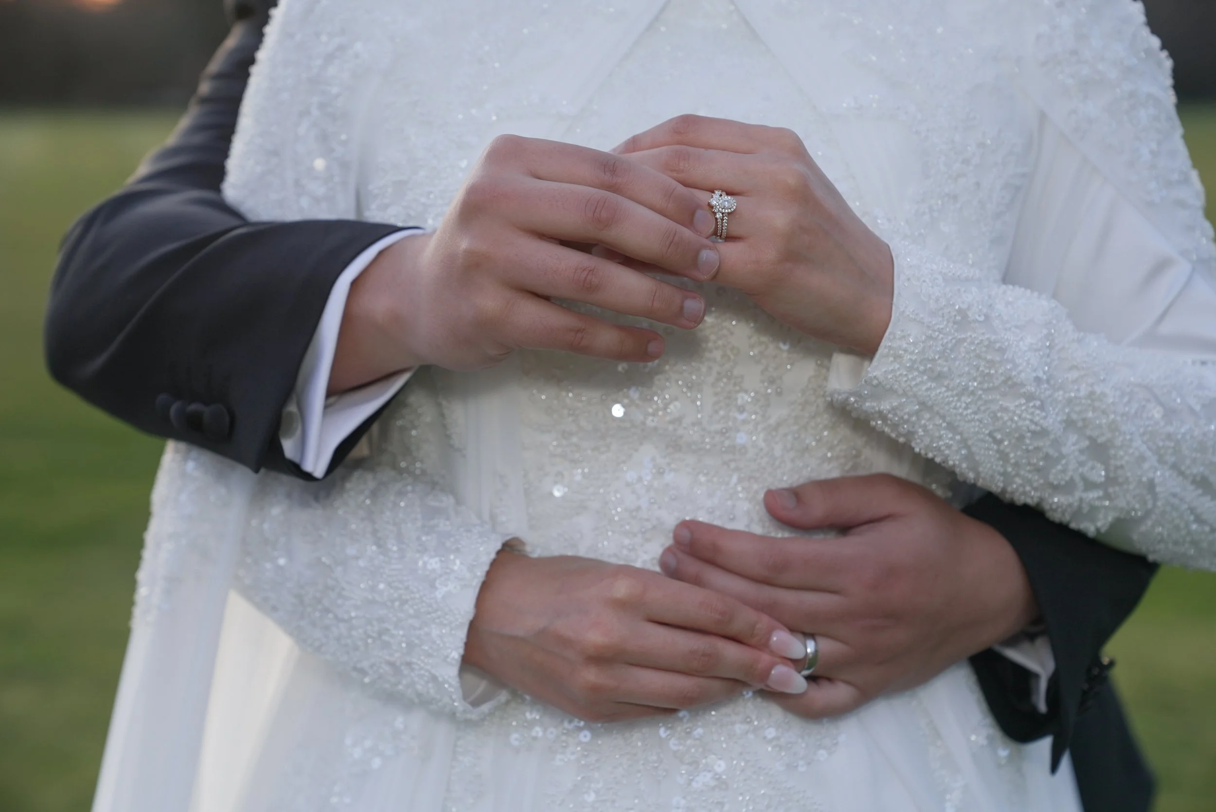 A bride and groom embrace, showing close-up of their hands with wedding rings. The bride's hand is on top, displaying a diamond engagement ring and wedding band, and the groom's hand is beneath, with a wedding band. The bride's dress is decorated wit