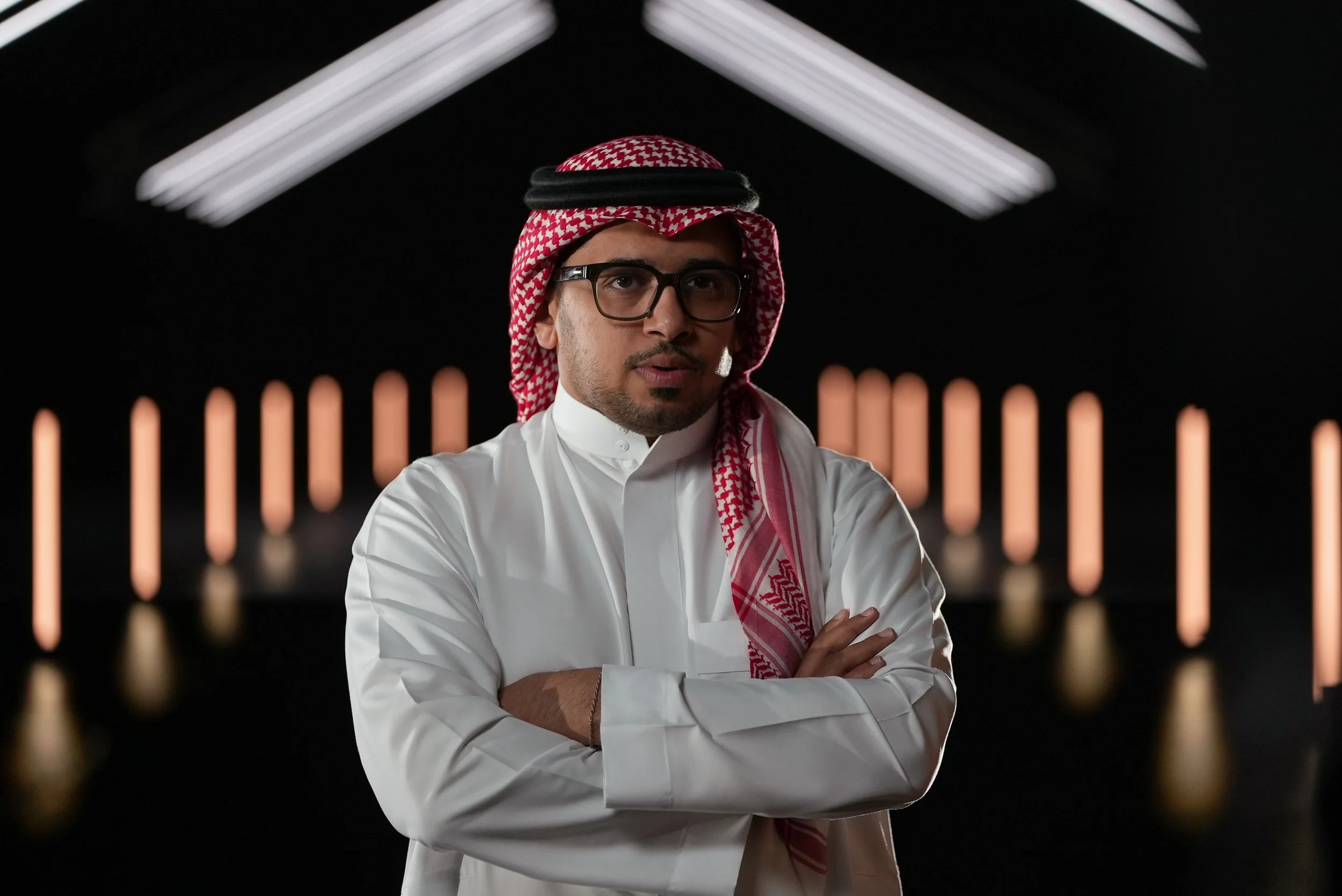 Man wearing traditional Middle Eastern attire, including a white thobe and red-and-white checkered keffiyeh, standing with arms crossed against a dark background with illuminated vertical lights.