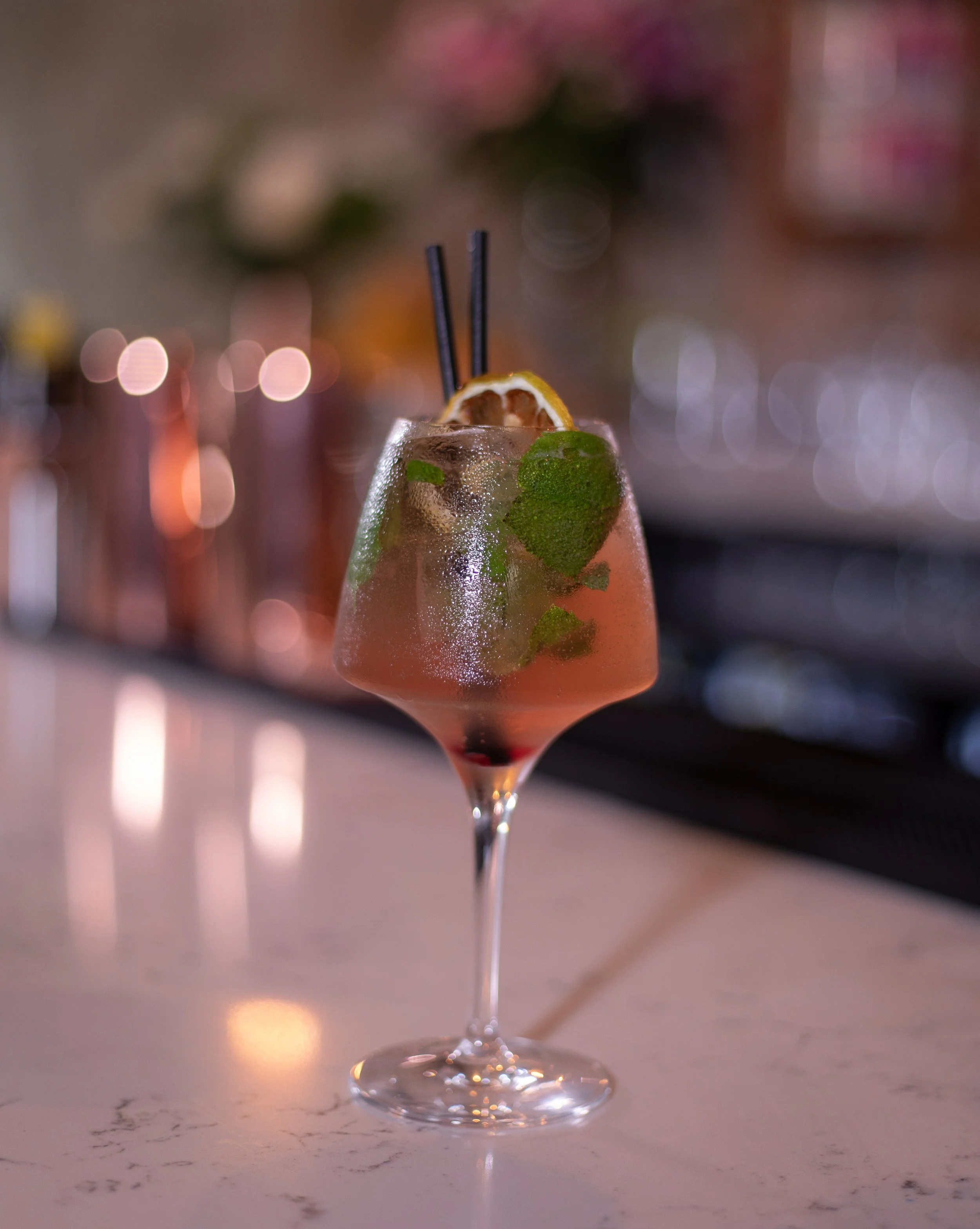 A pink cocktail with mint leaves, lemon slice, two black straws in a tall glass on a bar counter.