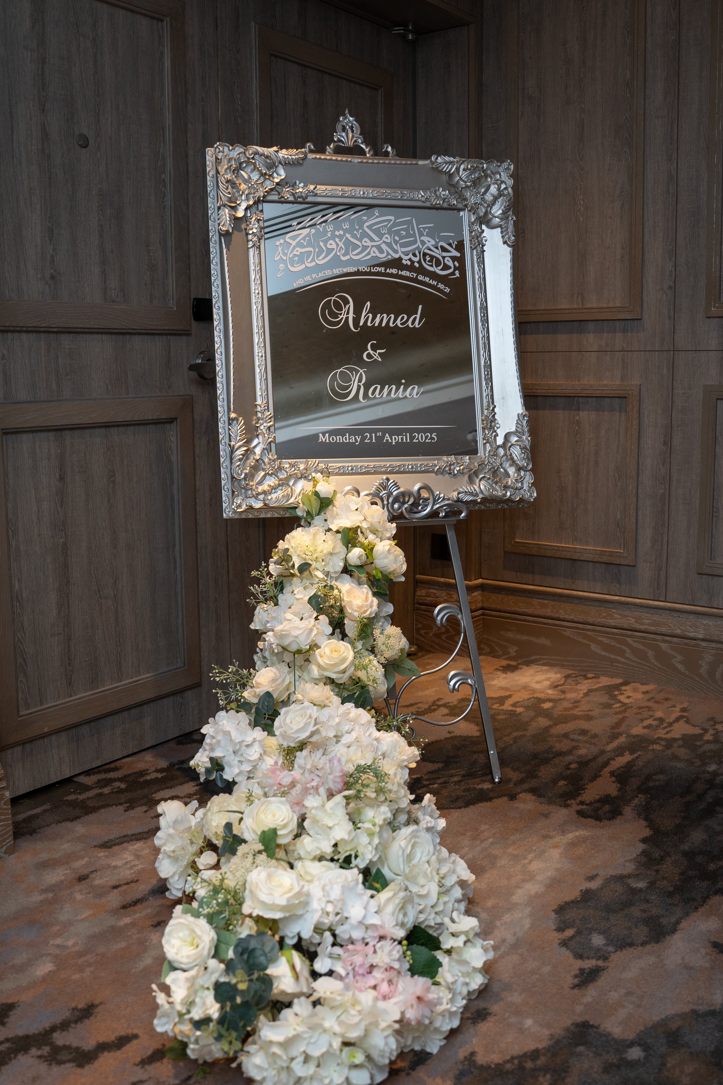 Wedding sign with names Ahmed and Rania, date April 21, 2025, on easel, decorated with white and light pink flowers, in a wood-paneled room.