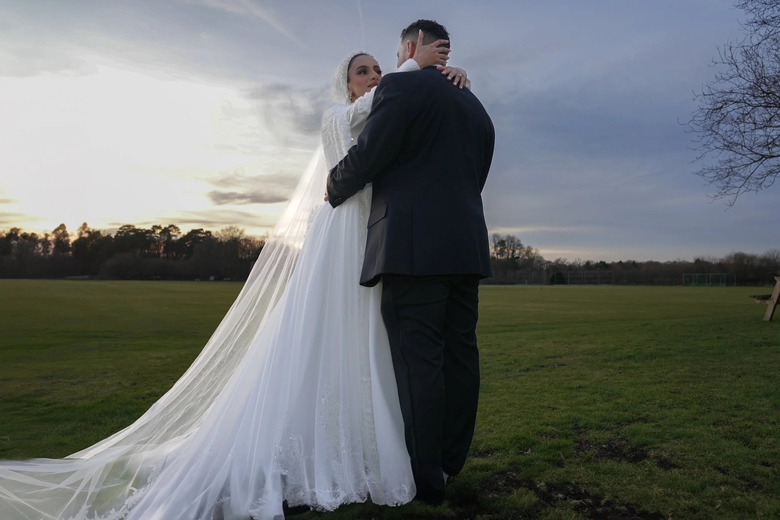 Bride and groom embracing outdoors during sunset, bride in a white wedding gown with a long veil, groom in a dark suit, open field and trees in the background.