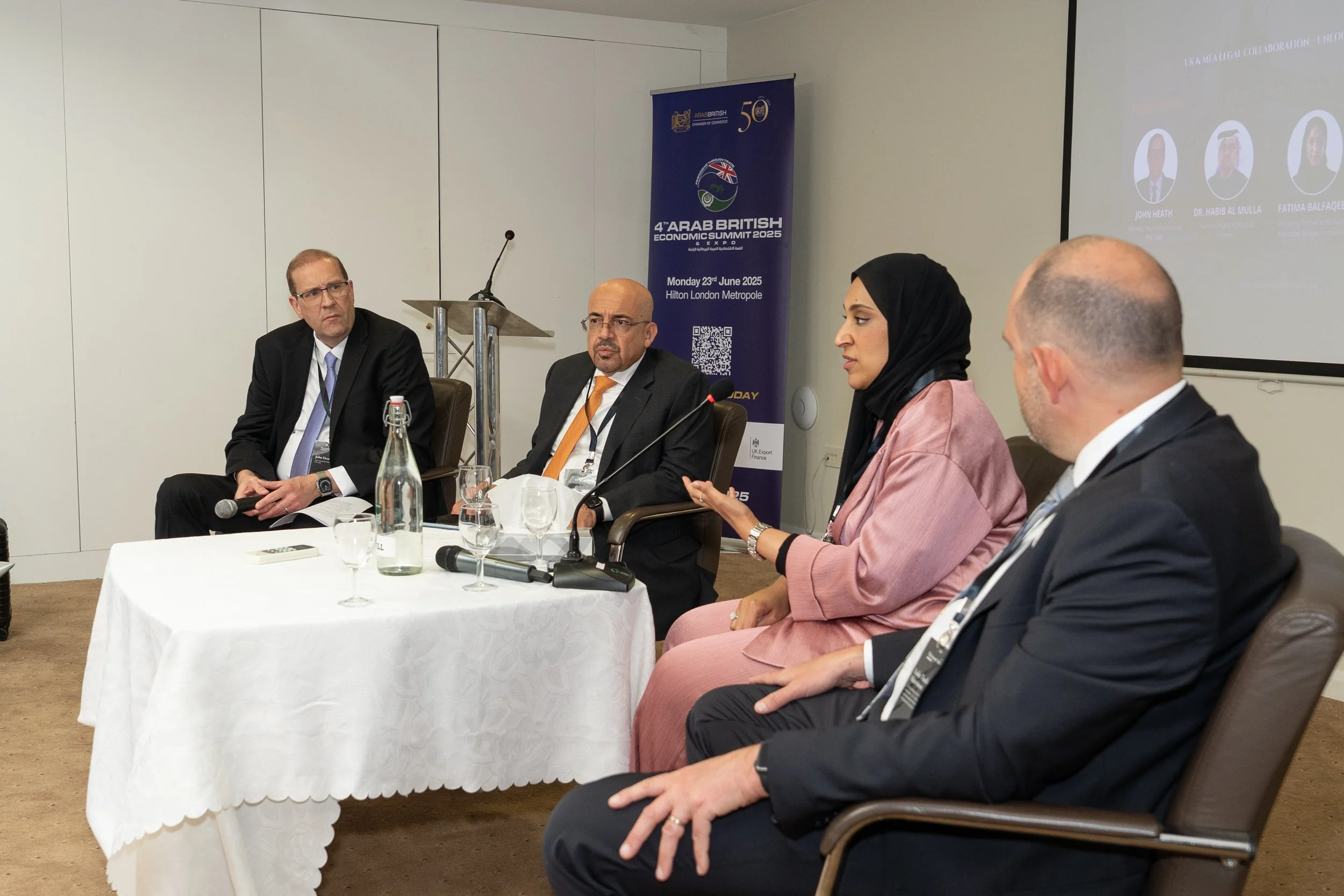 A panel of four people sitting at a conference table during a discussion at the Arab British Economic Summit 2025, held in a hotel conference room. There is a large screen on the wall showing the event's logo and speakers' photos.