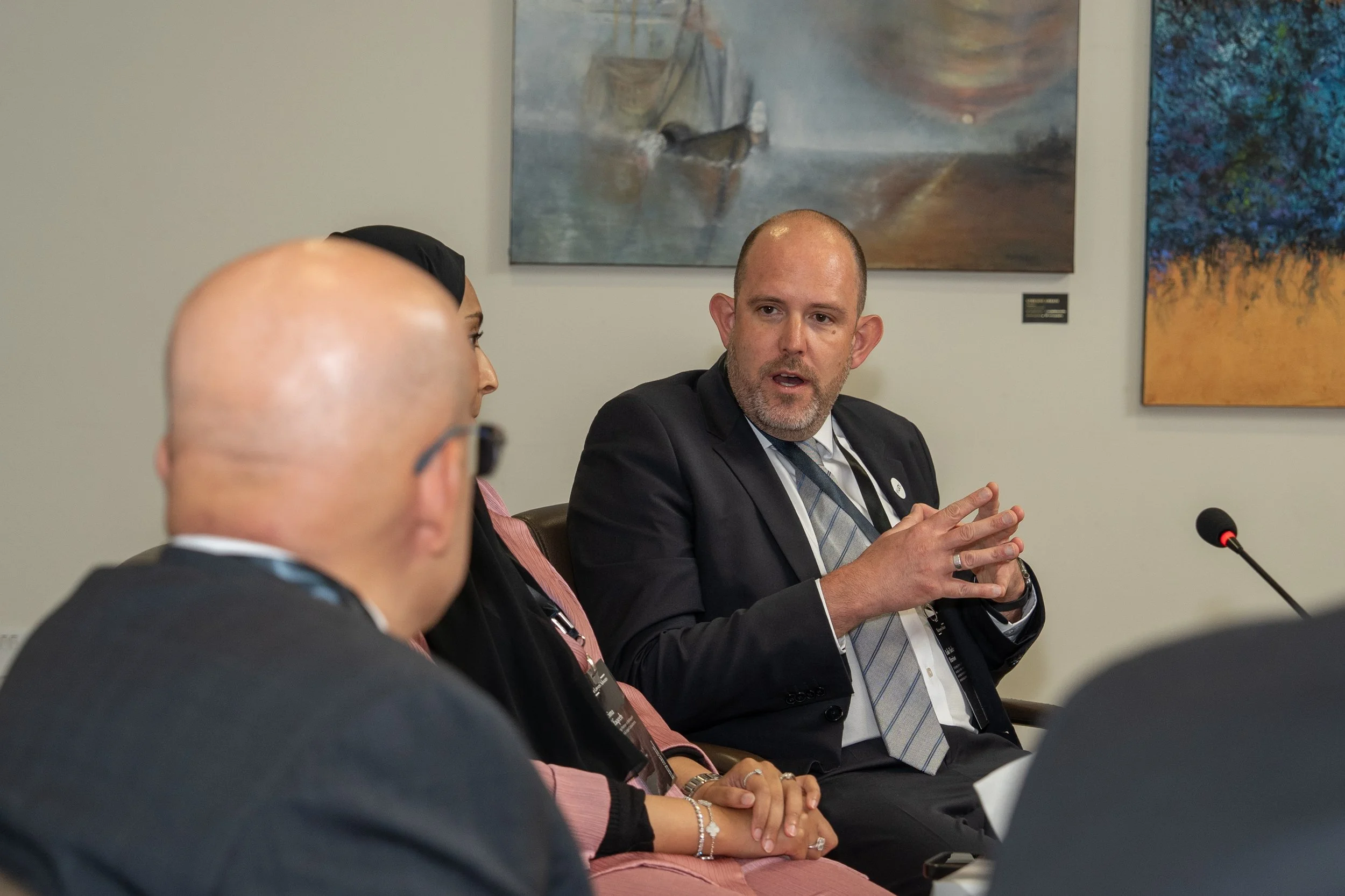 A group of three people sitting at a table during a discussion or meeting, with one man speaking and gesturing with his hands. The background shows artwork on the wall.