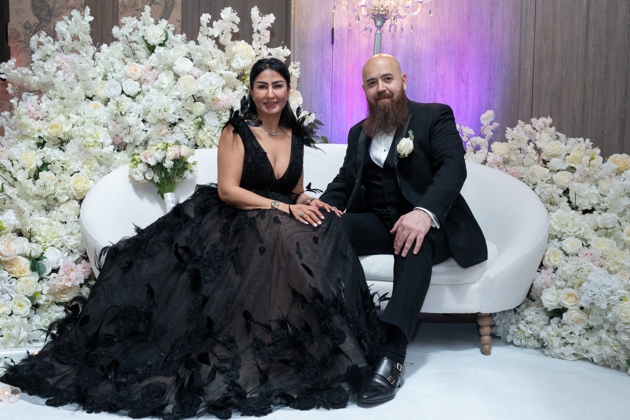 A woman in a black gown and a man in a tuxedo sitting on a white sofa surrounded by white floral decorations at a formal event.