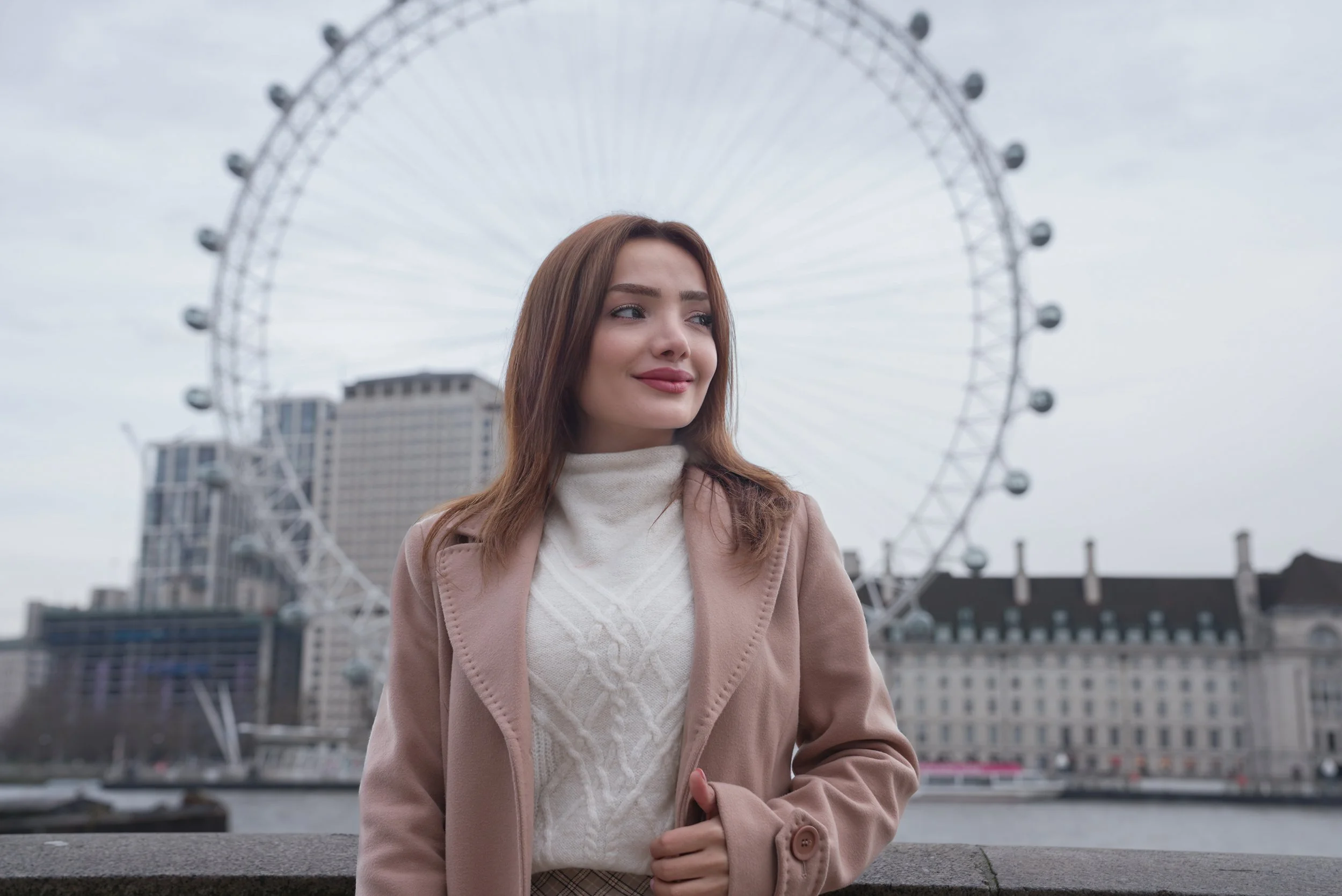 Young woman standing outdoors near river, with London Eye Ferris wheel and city buildings in background on overcast day.