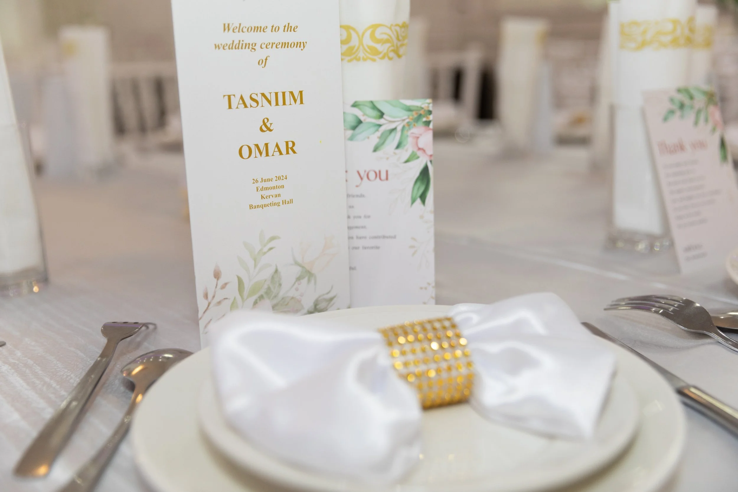 Wedding reception table setting with a white napkin and gold embellishment on a plate, space for utensils, and welcome cards for the wedding of Tasniim and Omar, dated June 26, 2024, in Edmonton at Kerven Banqueting Hall.