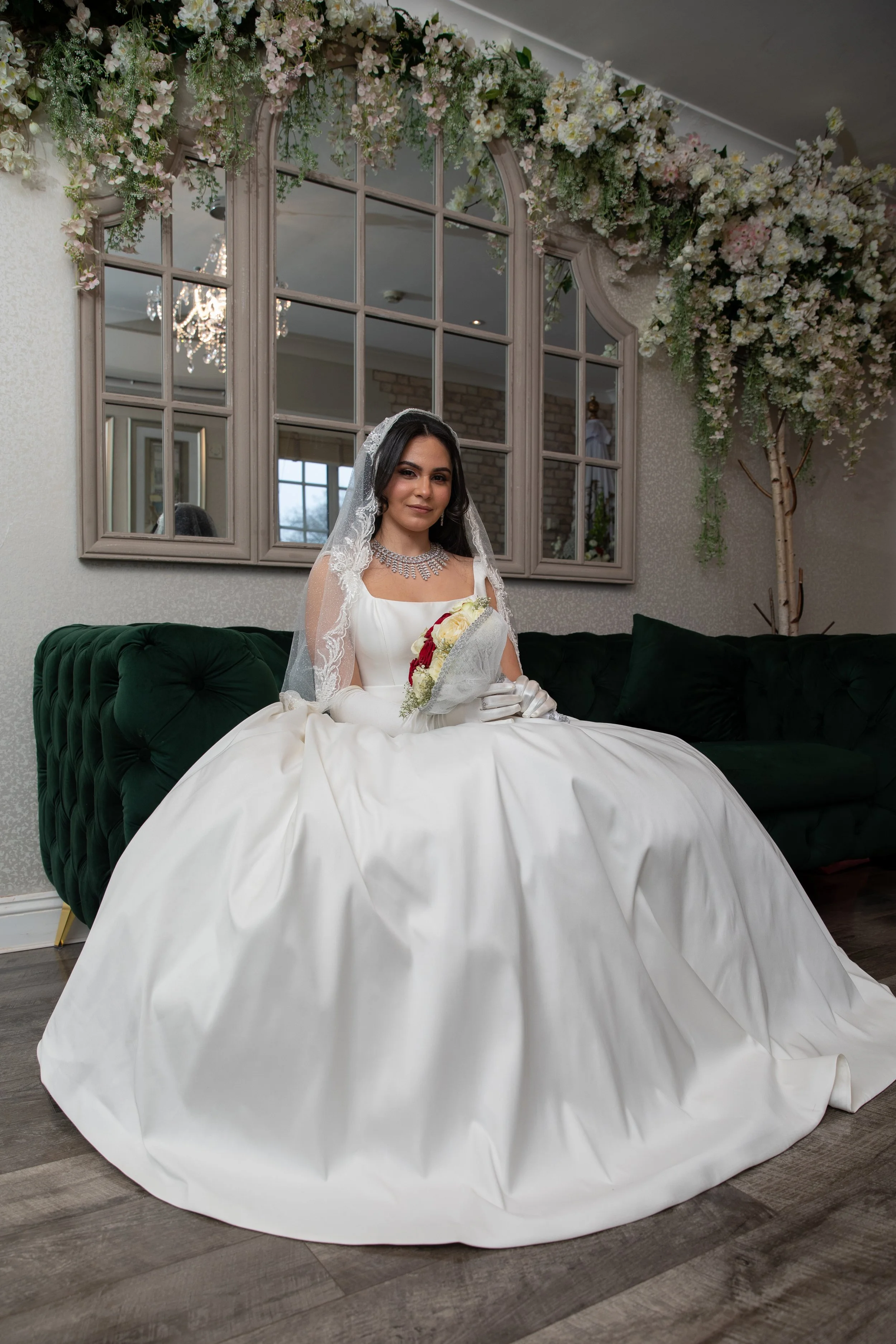 A bride sitting on a dark green velvet sofa, wearing a white wedding gown, veil, gloves, and holding a bouquet of white and red flowers, indoors decorated with flowers and a large mirror.