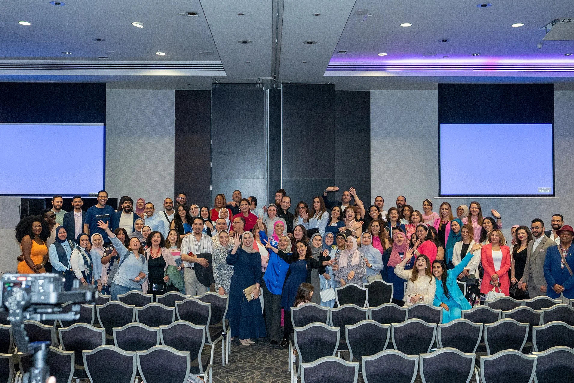 Group of diverse people gathered in a conference room, posing for a photo, with some people smiling and waving.
