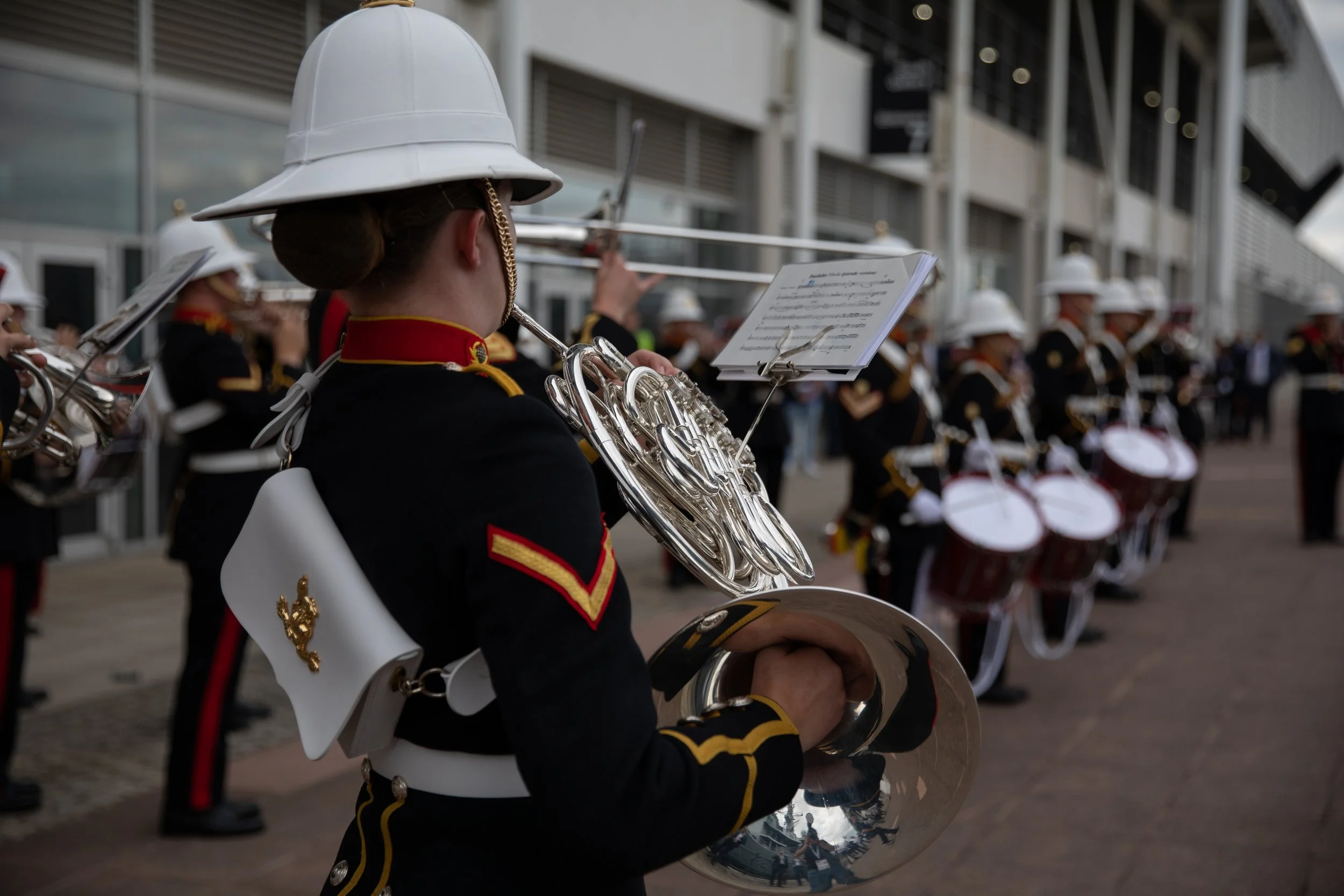 A marching band performing outdoors with members dressed in black uniforms with red and gold accents, wearing white helmets, playing various brass instruments, including a French horn and drums.