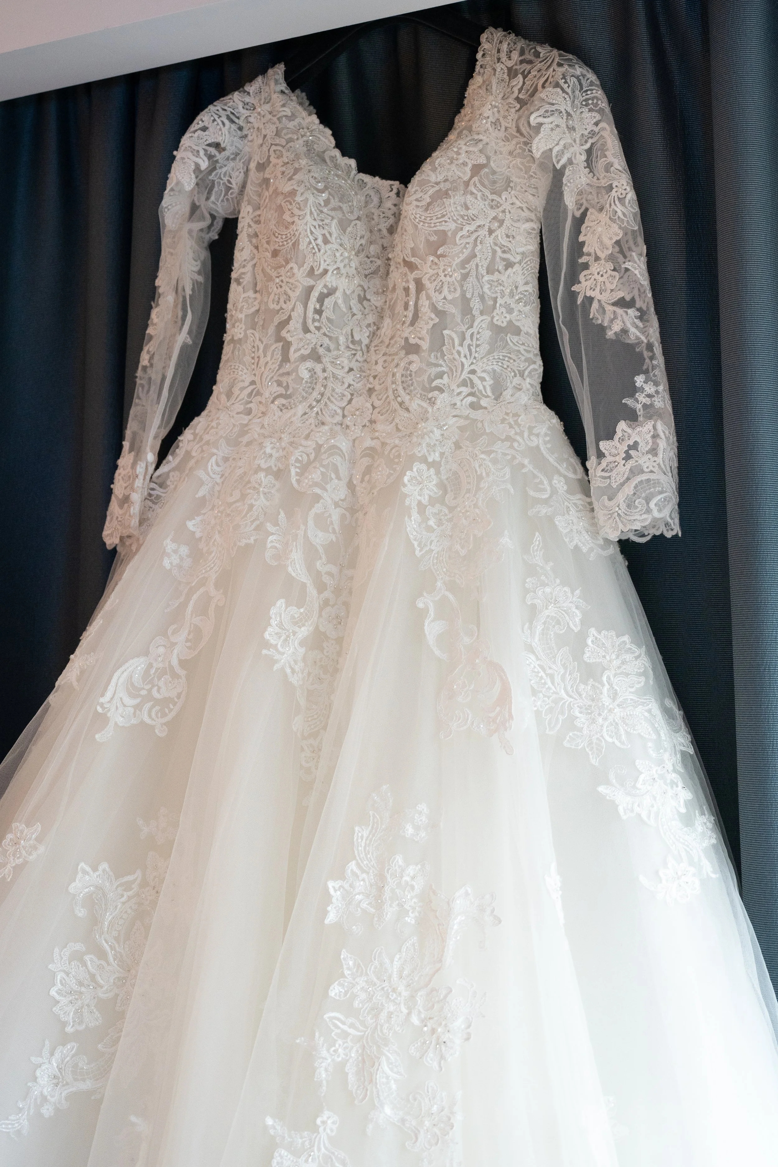 A wedding gown with intricate lace details hanging against a dark curtain.