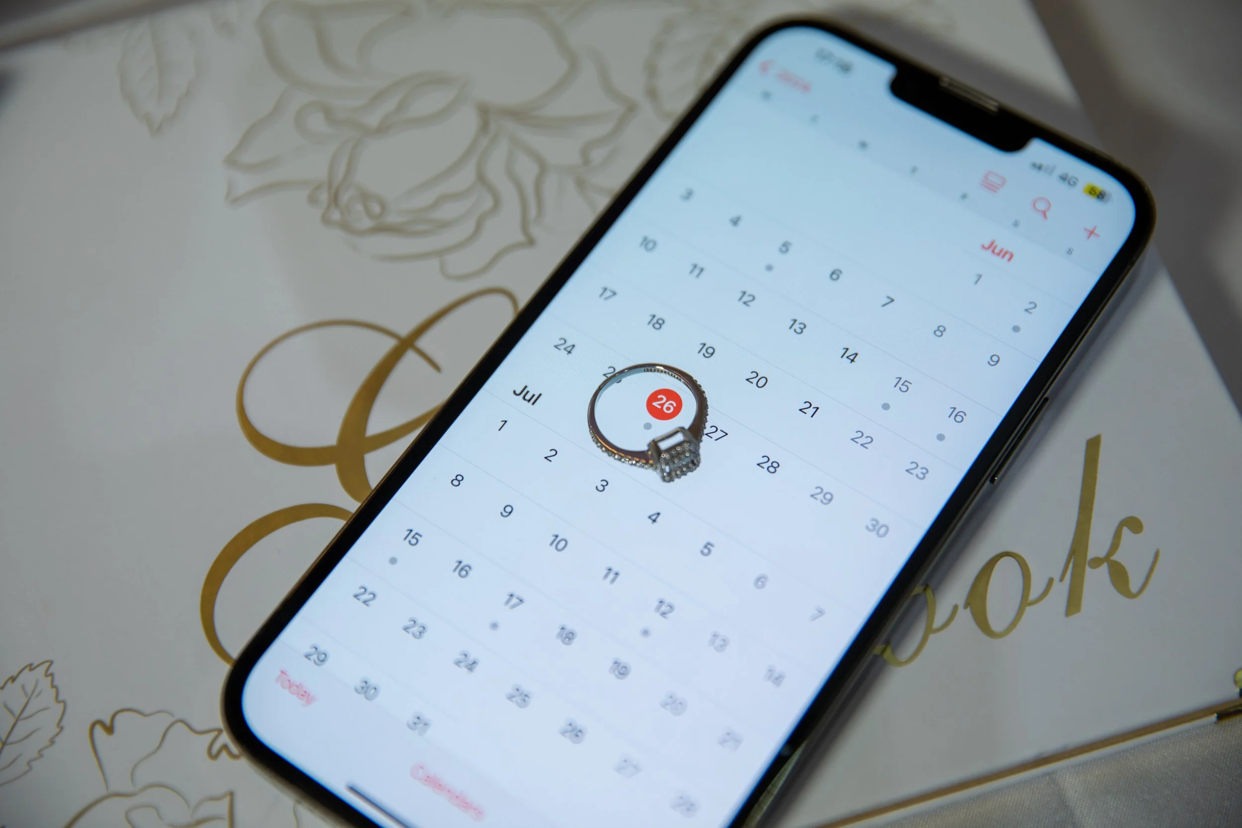 A smartphone displaying a calendar app with the date July 26 highlighted, placed on top of a book with gold and black lettering and floral design.