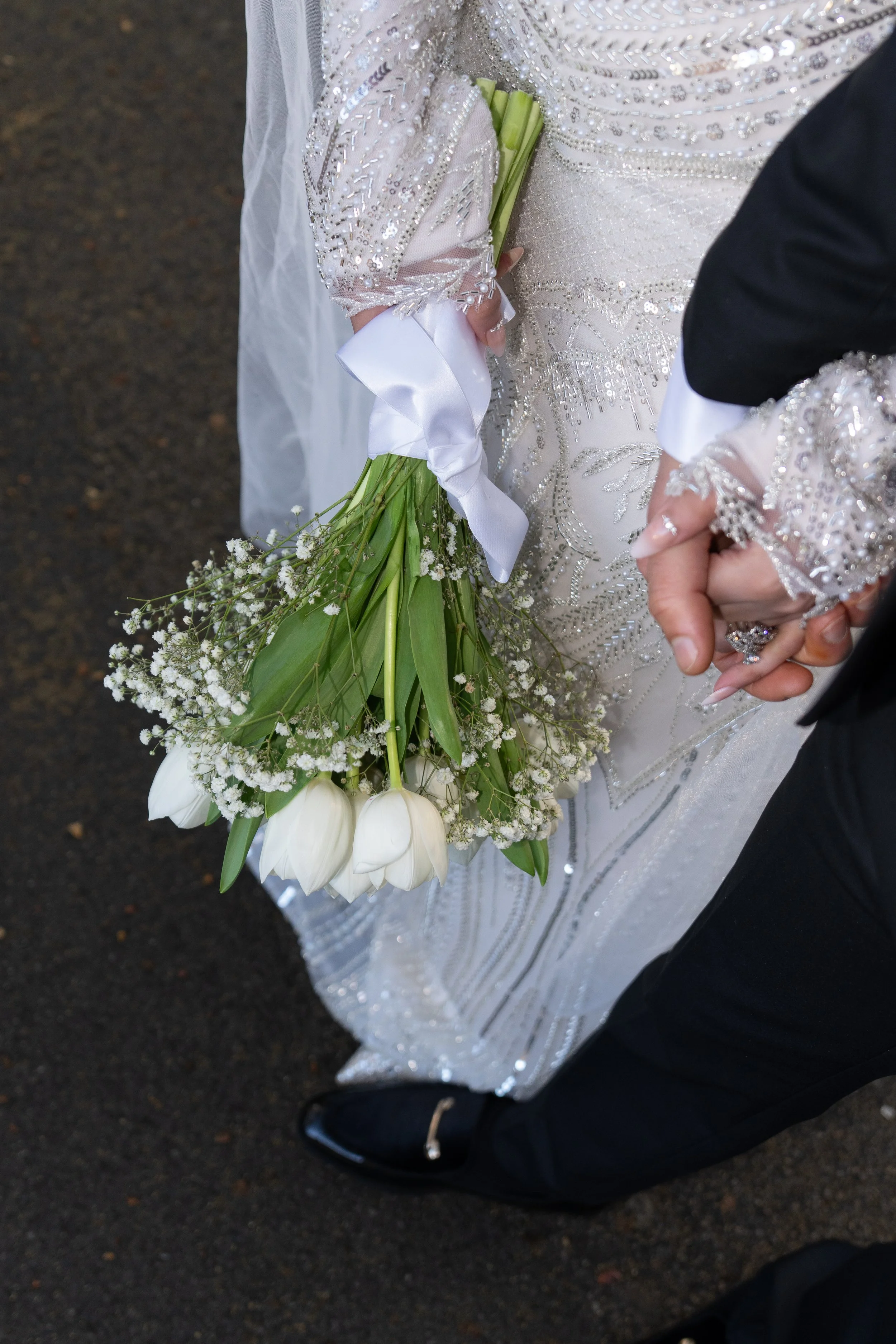 A bride in a beaded wedding dress holding a bouquet of white tulips and baby's breath, walking with a groom at a wedding.