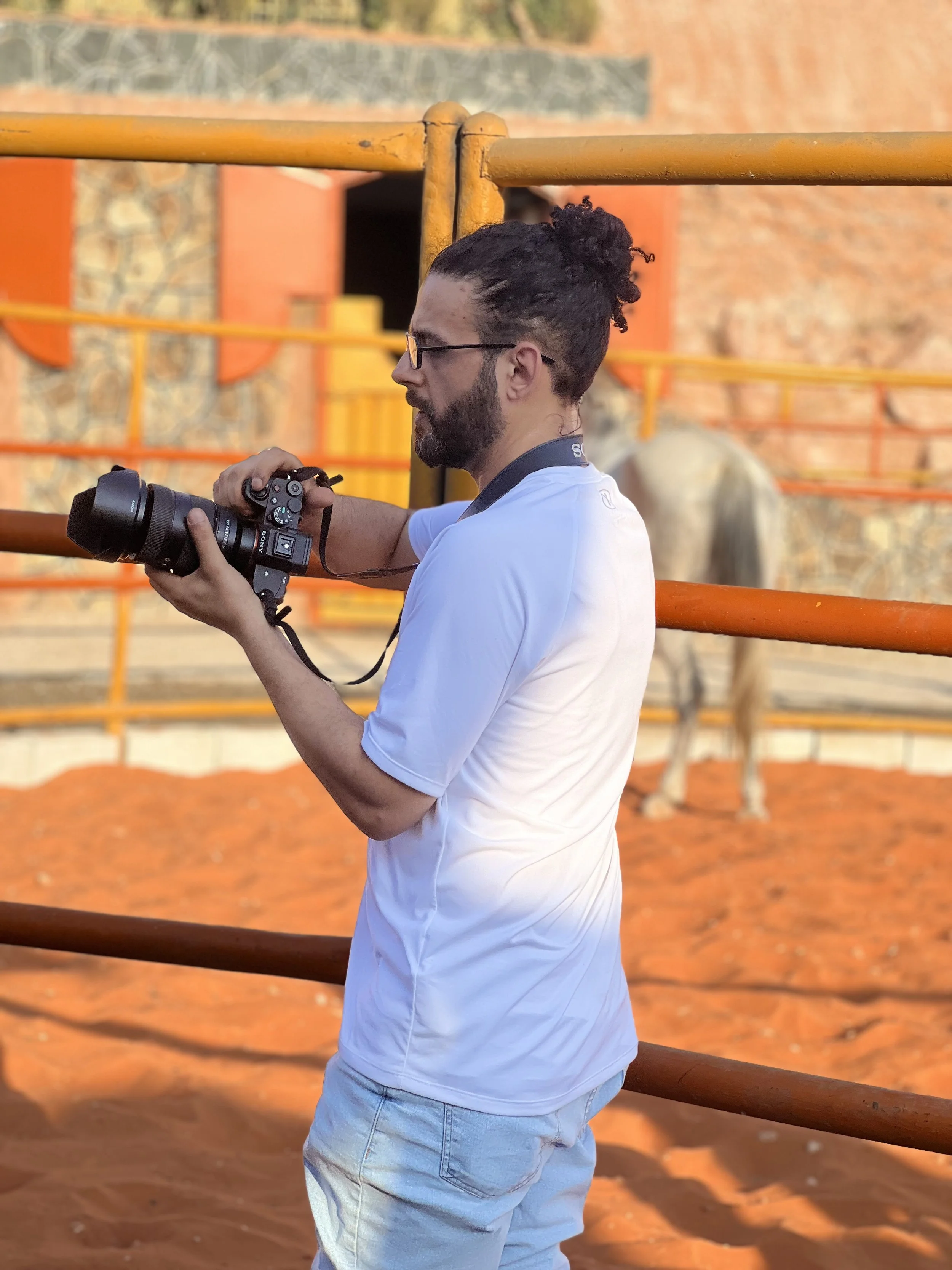 A man with glasses and long curly hair tied back in a bun, wearing a white shirt, is holding a camera and looking at its screen. He is standing outdoors near orange safety railings, with an elephant in the background.