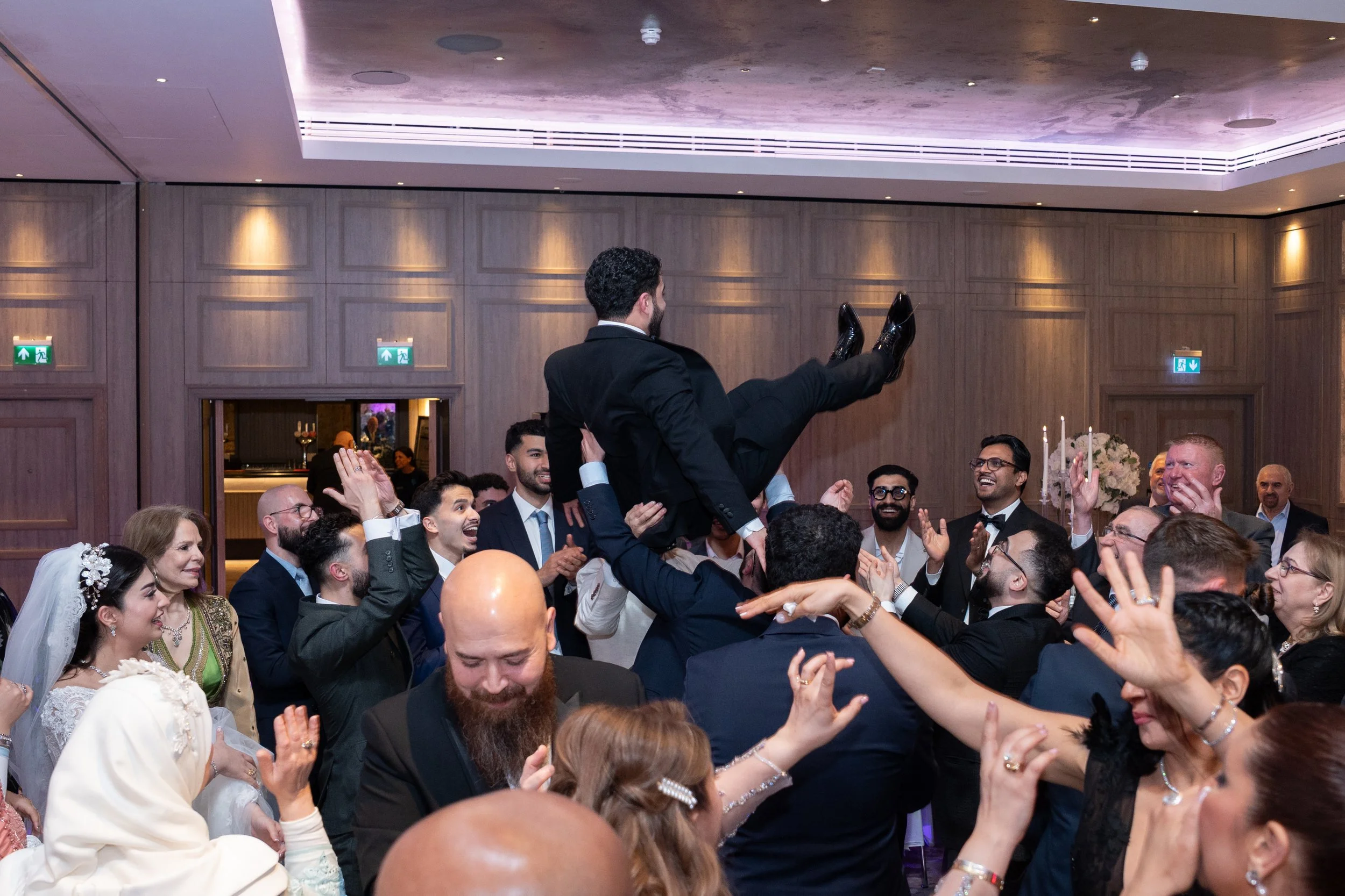 People at a wedding celebration lift a man in a black suit and dress shoes in a chair above their heads, surrounded by smiling guests.