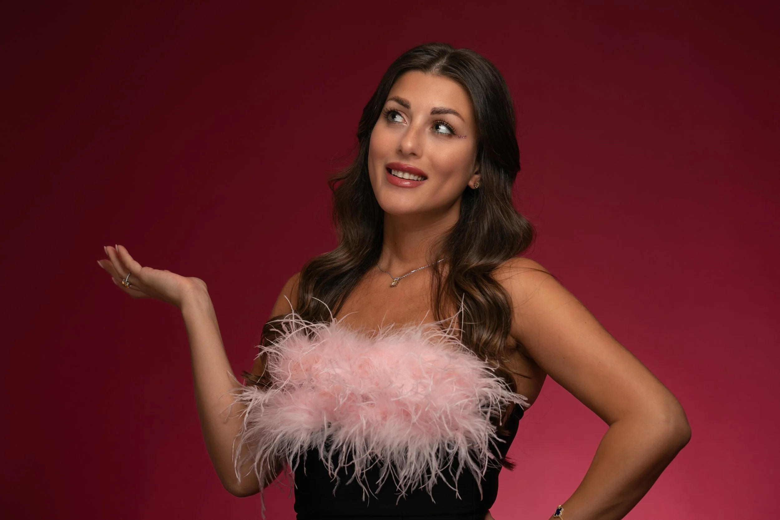 Woman with long brown hair wearing a black dress with pink feathered detail on the chest, posing with her left hand on her hip and right hand raised, against a red gradient background.