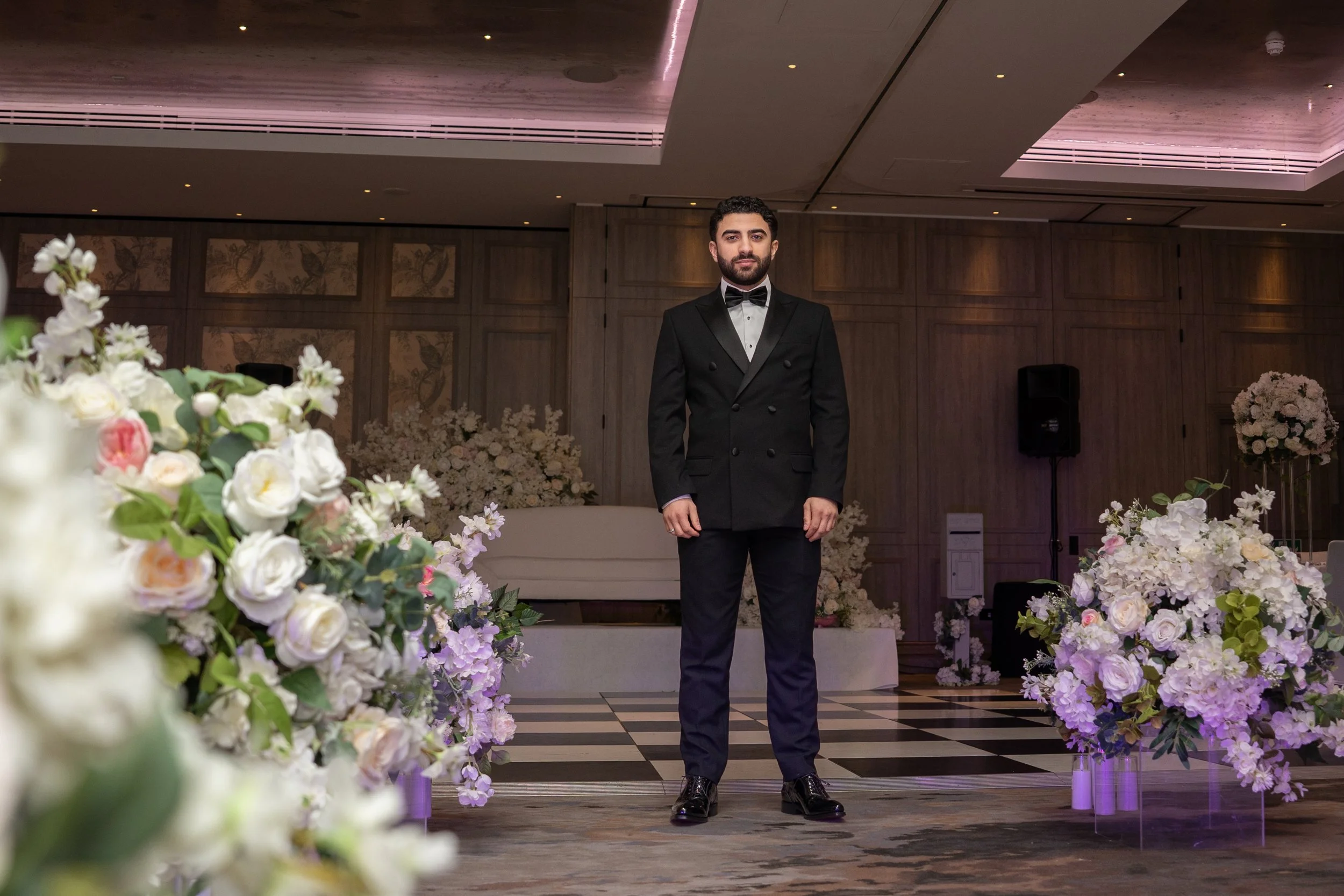 A man dressed in a black tuxedo with a bow tie standing in a decorated indoor setting with flowers and flowers arrangements in the background.