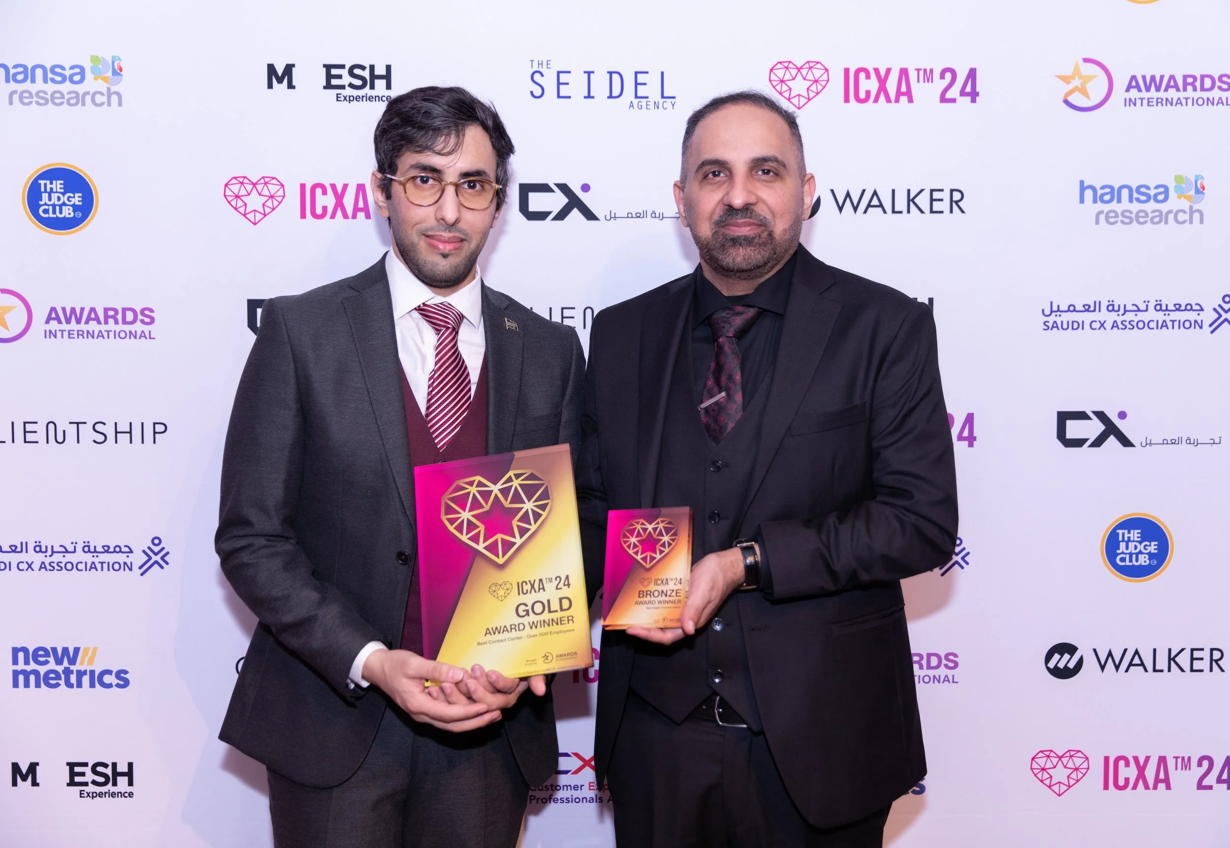 Two men in suits standing in front of a backdrop, holding awards plaques at an event celebrating ICXA 24 awards, with various sponsor logos on the backdrop.
