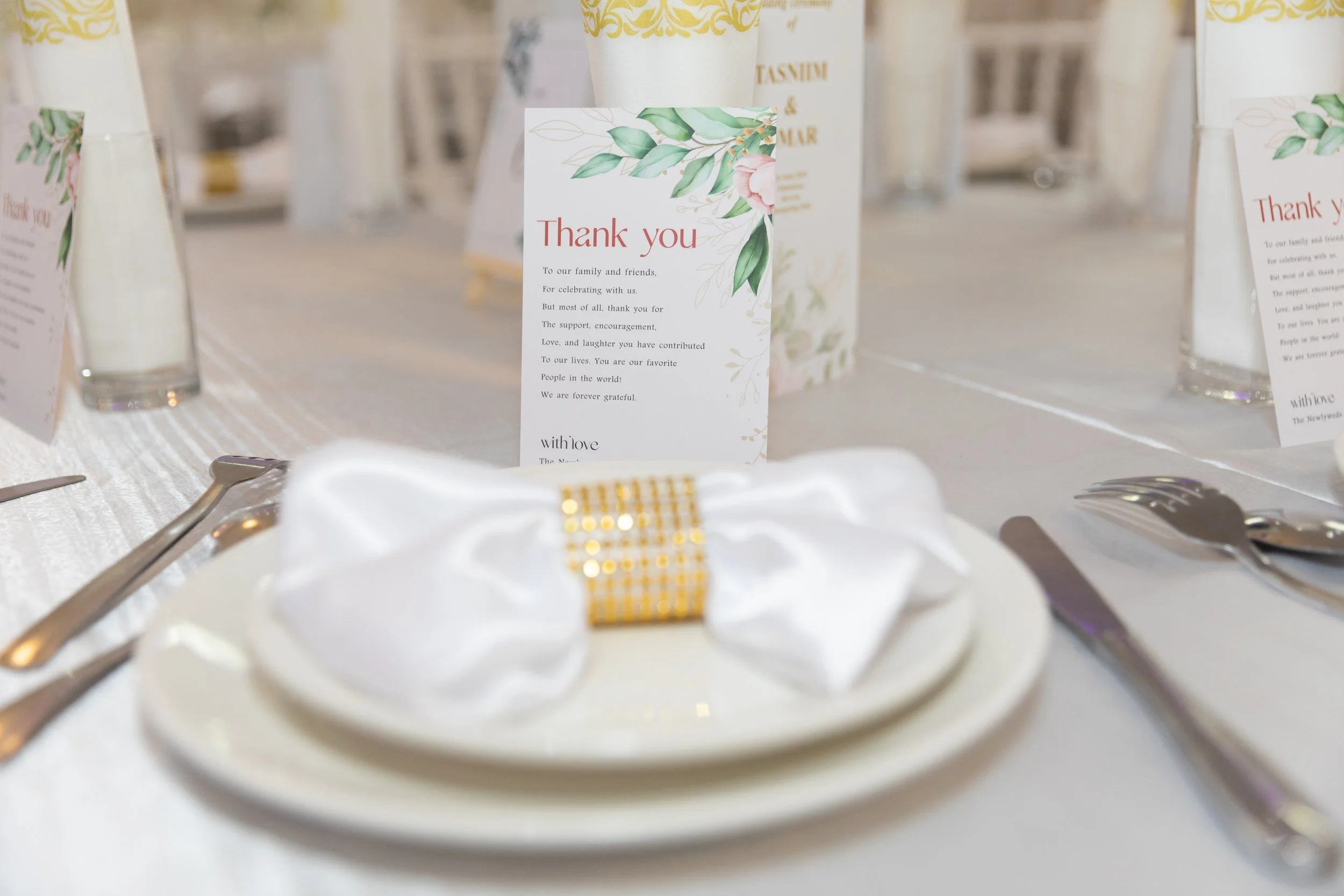 A wedding reception table setting featuring a white napkin wrapped with a gold band on a white plate, silverware including a knife and fork, and a 'Thank you' card with floral design in the background.