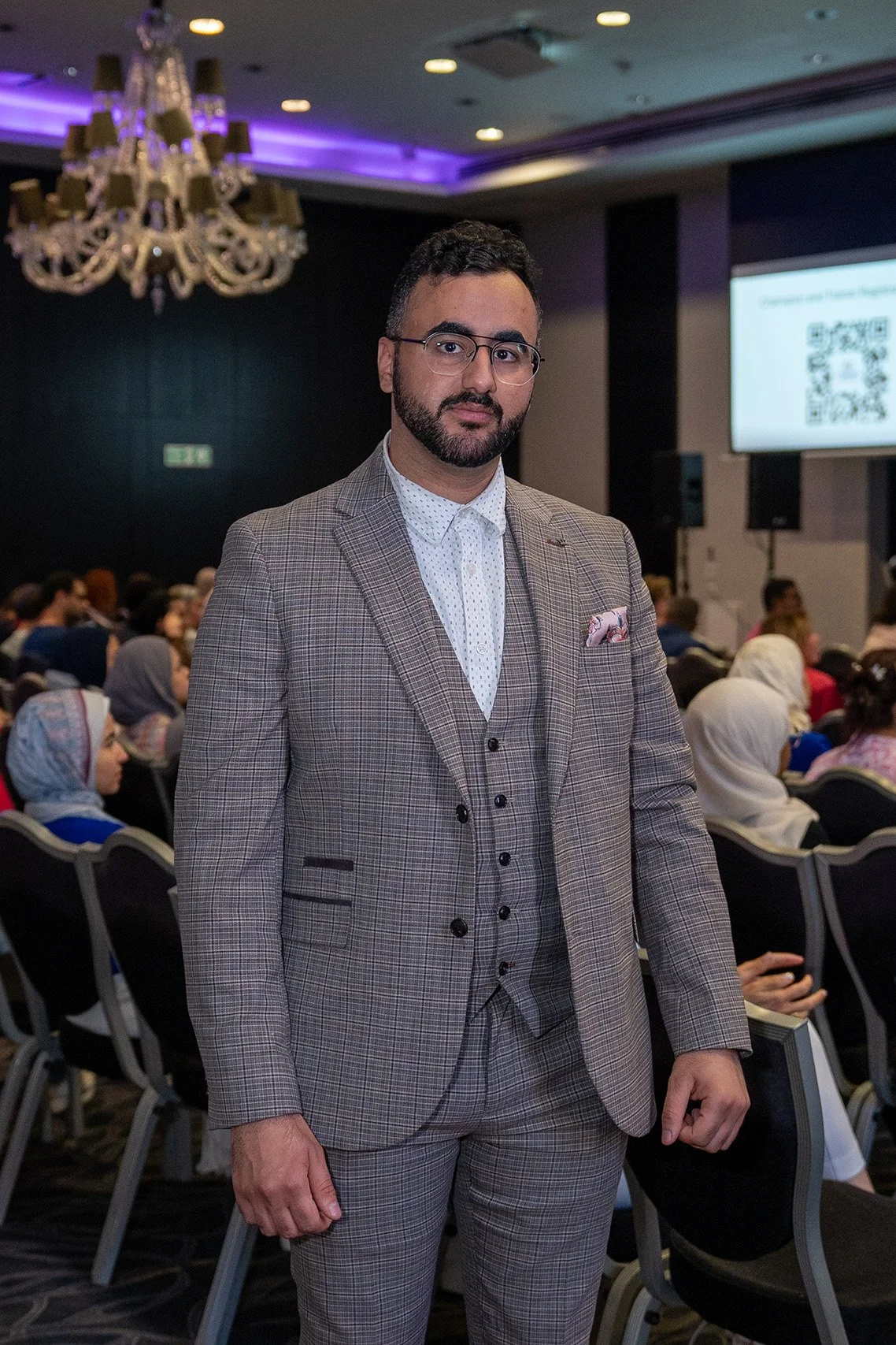 Man in a plaid suit standing in a conference room with attendees and a large screen in the background.