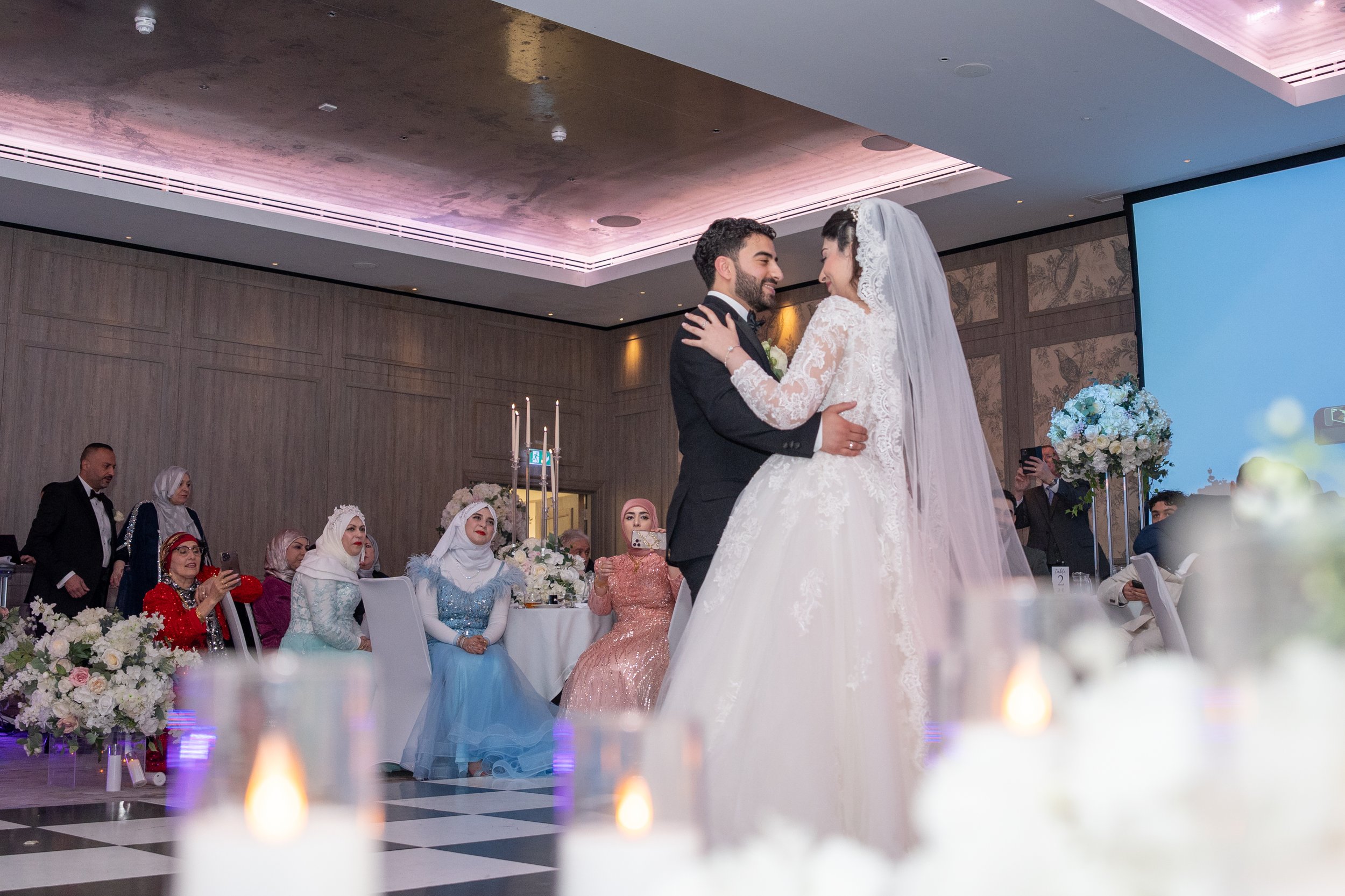 A bride and groom share a dance at their wedding reception, surrounded by guests seated at decorated tables with floral arrangements and candles, in a large decorated banquet hall.