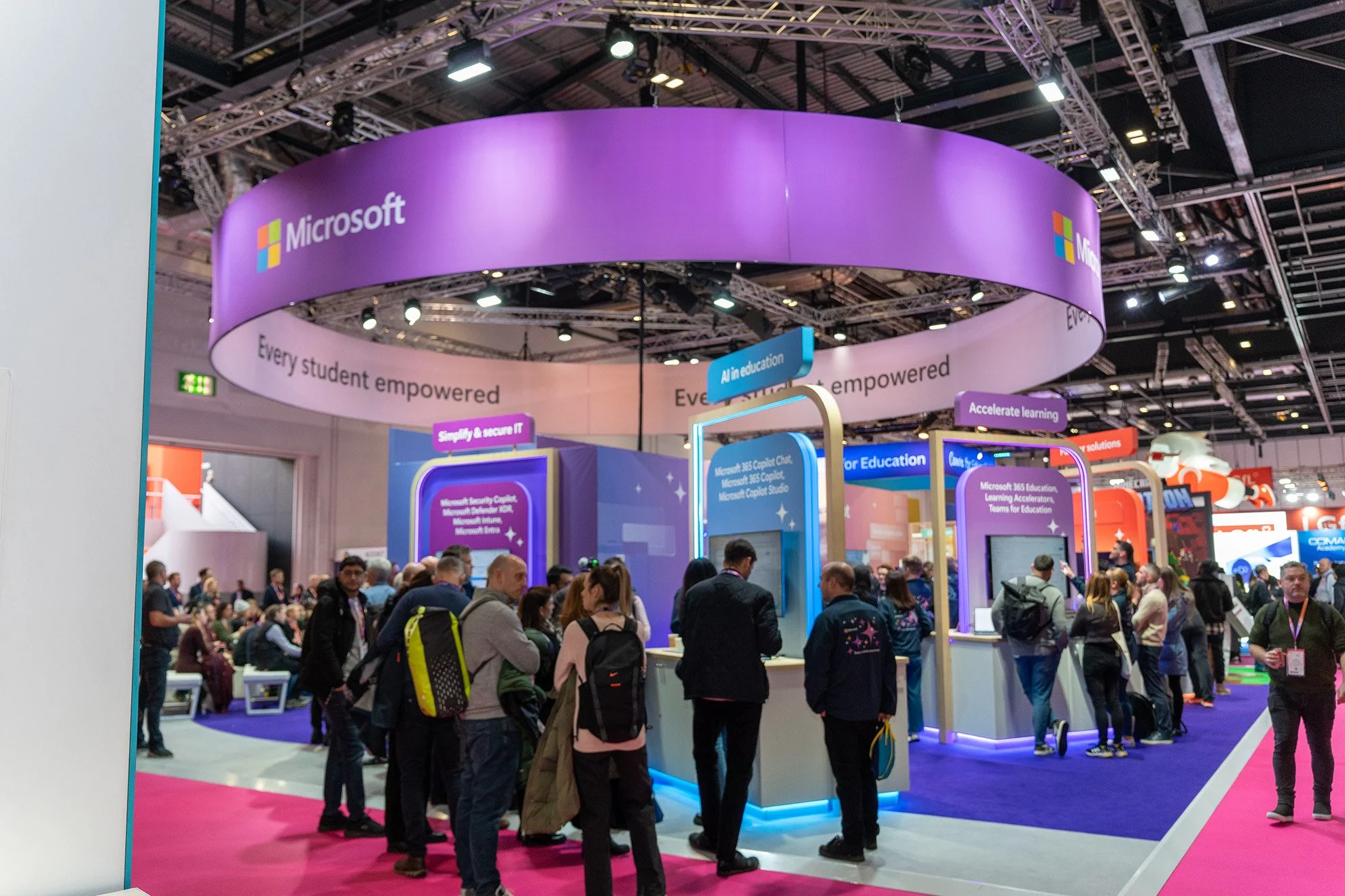 Microsoft booth at a technology conference with a large purple circular overhead sign displaying the Microsoft logo. The booth features various presentation stations with digital screens and signage promoting AI in education and secure IT solutions. 