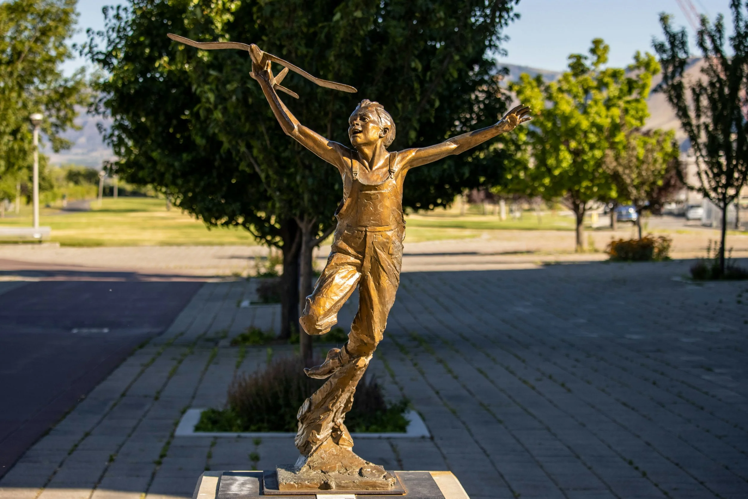 Bronze statue of a boy flying a paper airplane on a park sidewalk with trees and mountains in the background.
