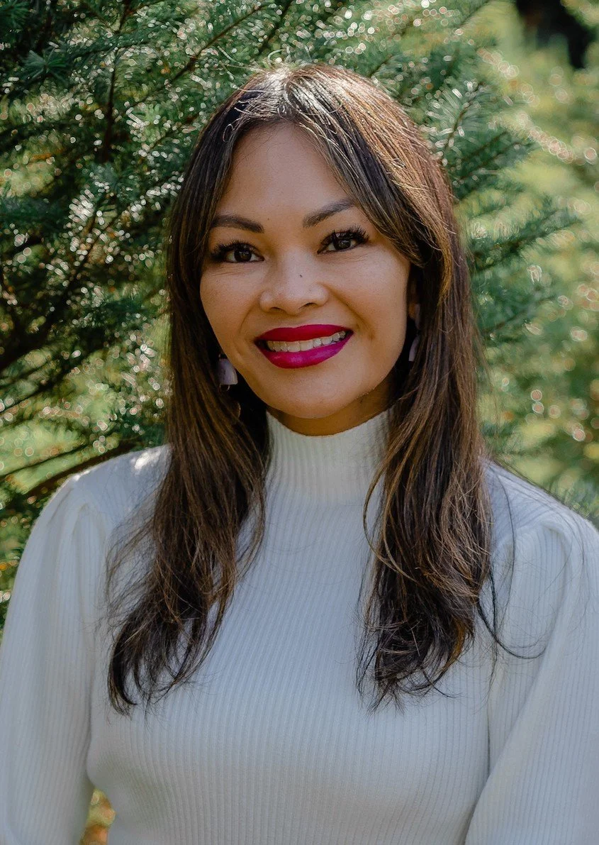 A woman with long brown hair and red lipstick smiling outdoors with green foliage in the background, wearing a white ribbed turtleneck sweater.
