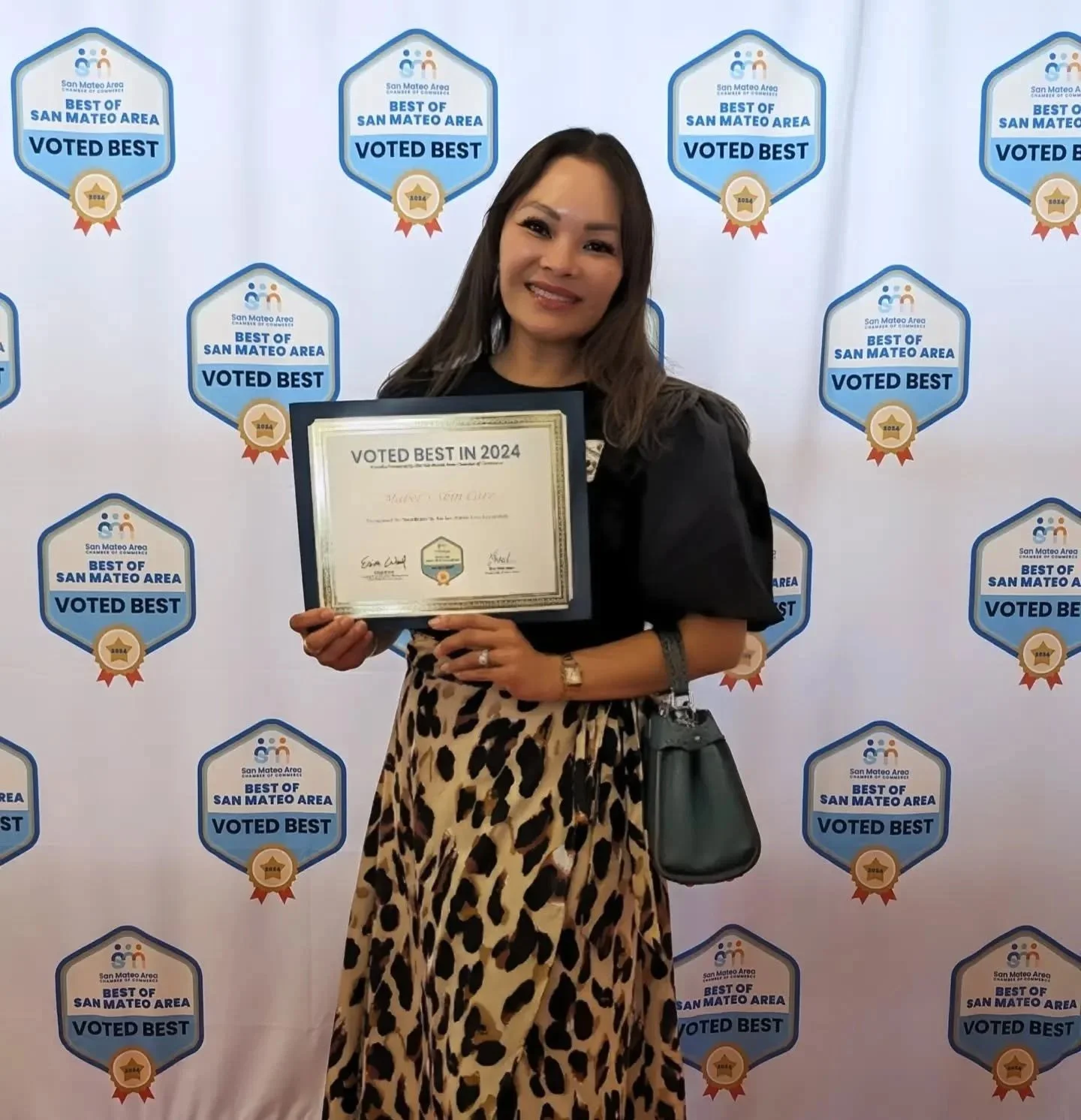 A woman standing in front of a backdrop with multiple 'Voted Best' badges from the San Mateo Area. She is holding a framed certificate that reads 'Voted Best in 2024.' She is smiling and wearing a black top and a leopard print skirt, with a black handbag hanging on her wrist.